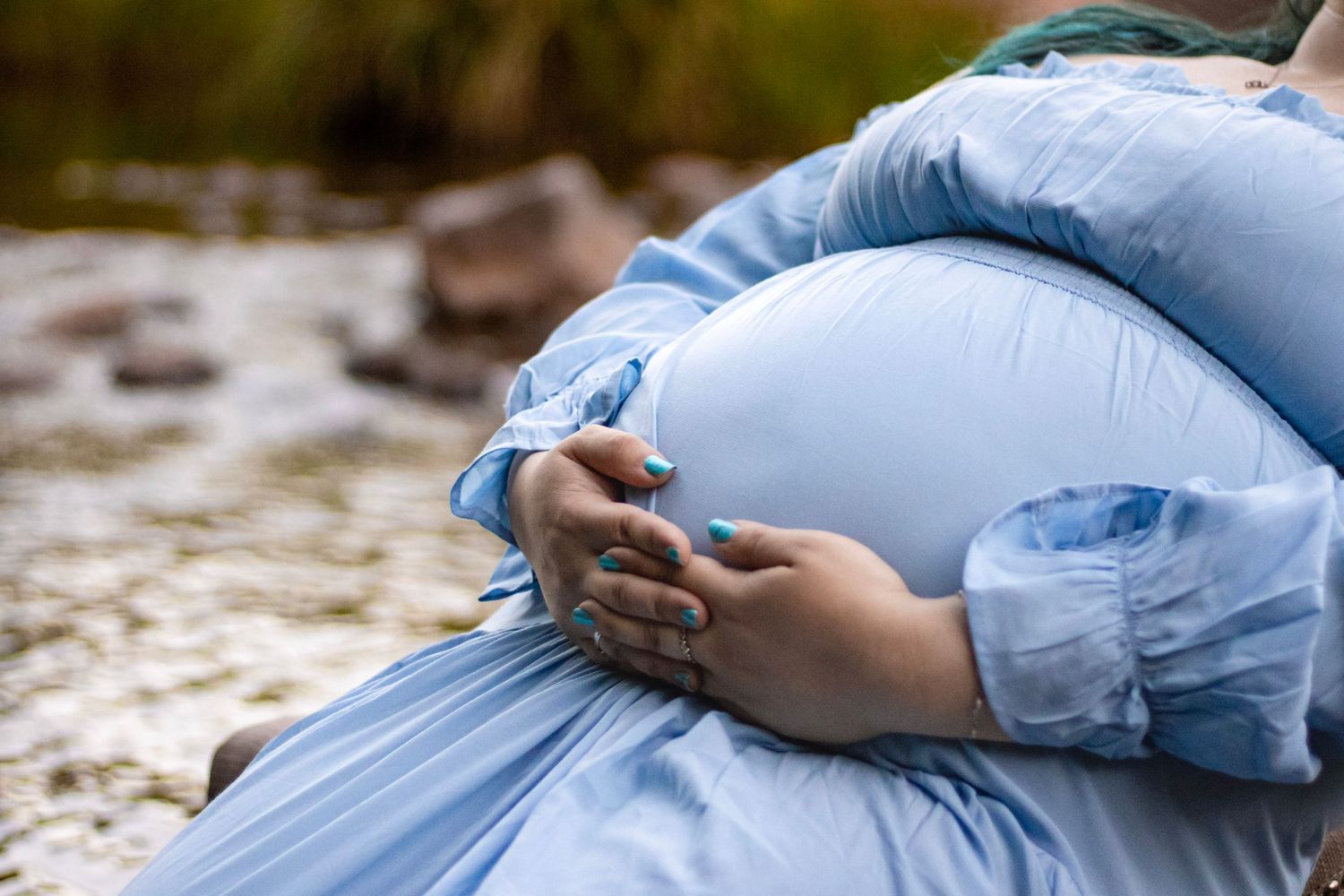 Close-up of pregnant belly cradled by hands in blue maternity dress outdoors.