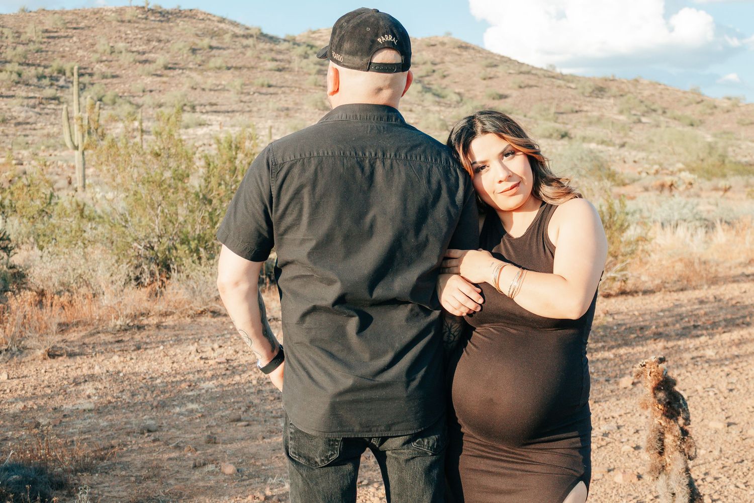 Pregnant woman in brown dress leans on man in black shirt during desert maternity photoshoot with cacti background.