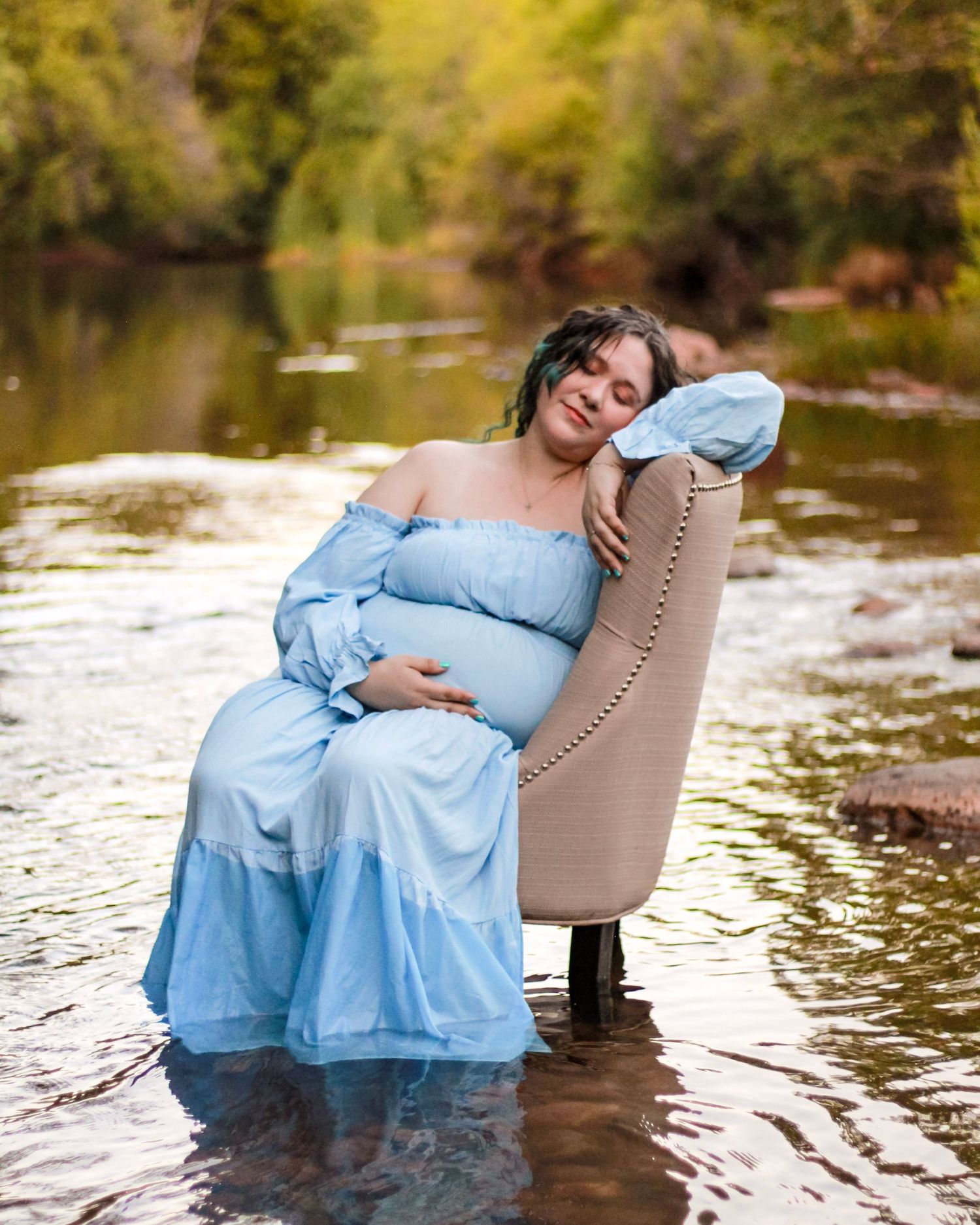 Pregnant woman in blue off-shoulder gown sitting in armchair in shallow river.