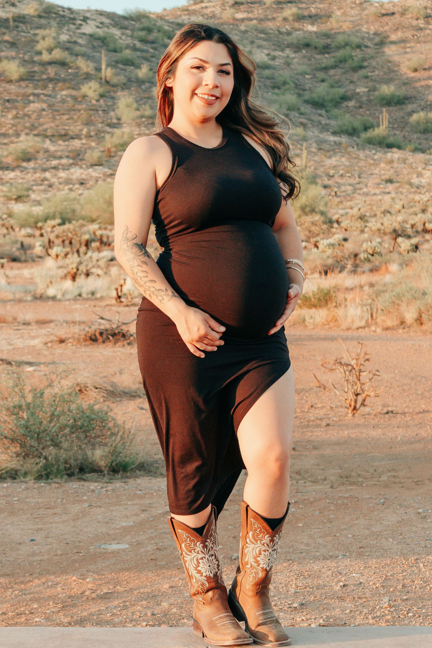 Pregnant woman in black dress and cowboy boots smiling outdoors in desert landscape.