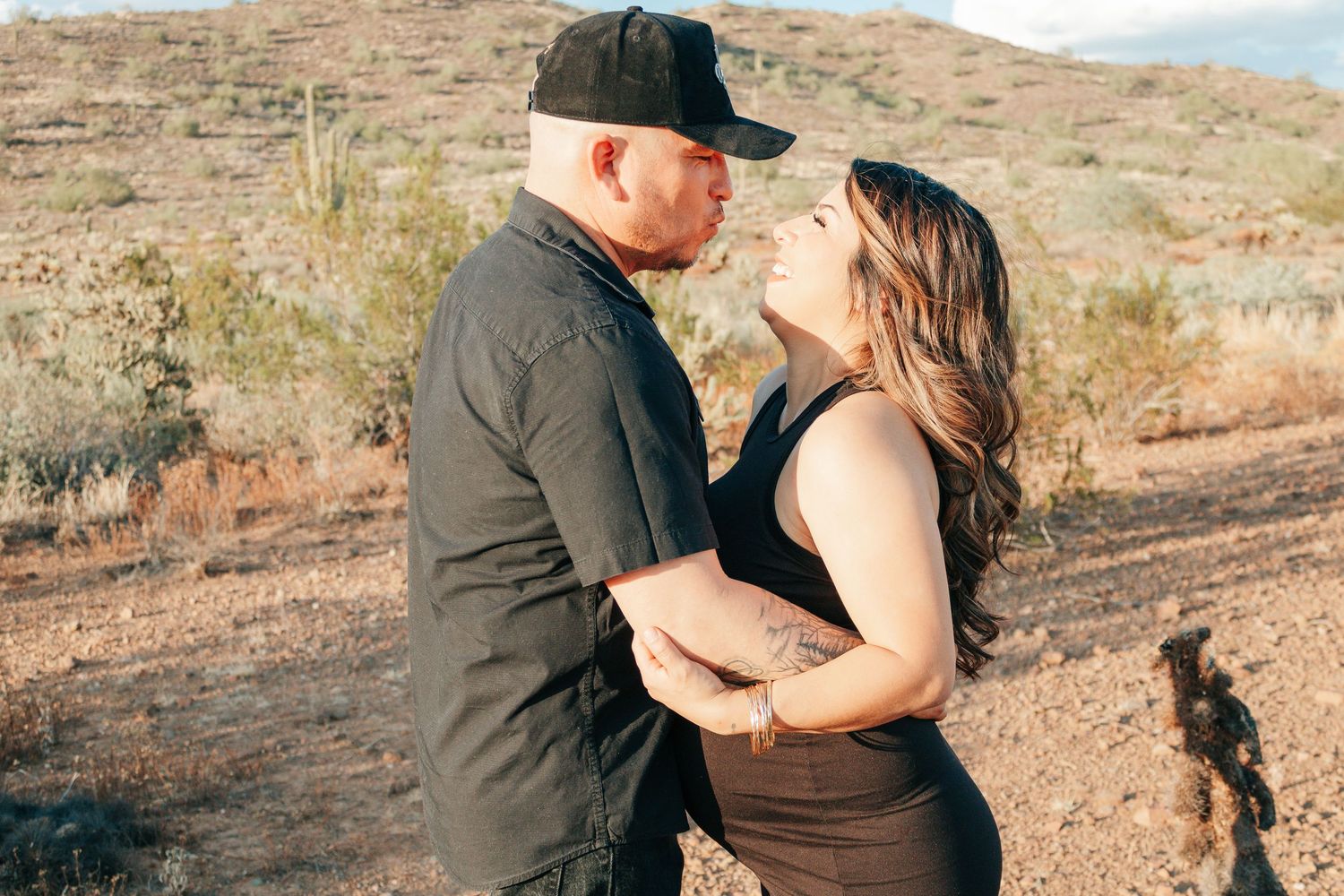 Couple sharing a romantic moment in a desert landscape with cacti and mountains in the background.