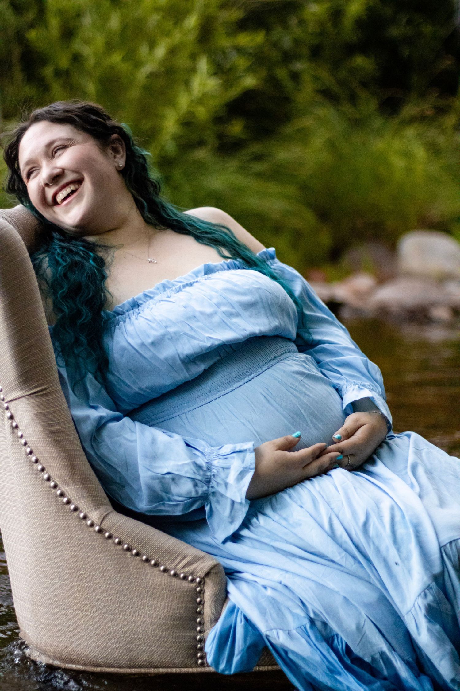Laughing pregnant woman with teal hair in blue gown seated in chair by river.