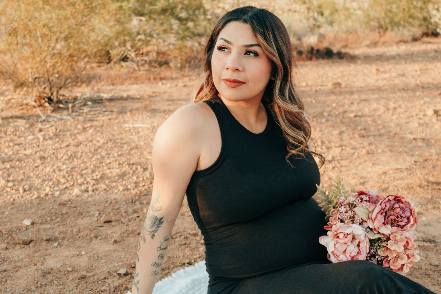 Woman in black dress holding pink flowers sits outdoors in desert landscape with tattoo on arm.
