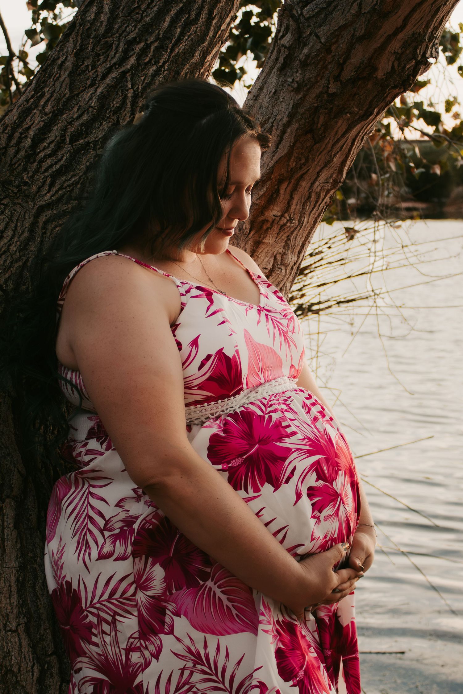 Pregnant woman in floral pink dress cradling baby bump, leaning against tree by water at golden hour.