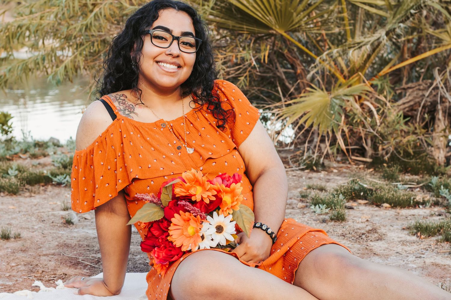 Young woman in orange outfit holding colorful flowers, smiling outdoors near palm trees and a serene waterway.