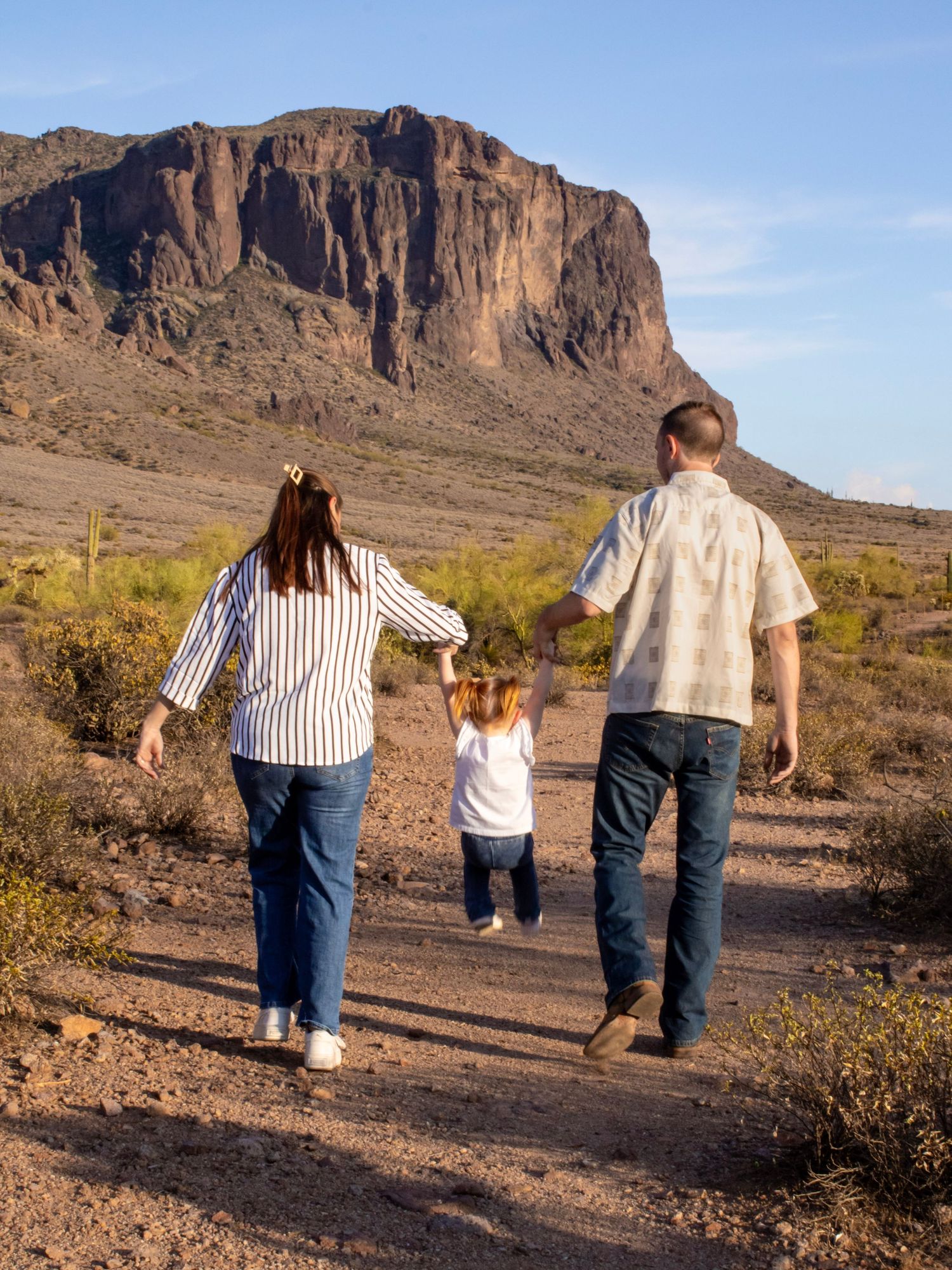 Family of three walking hand-in-hand on a desert trail with a dramatic rocky mountain in the background.