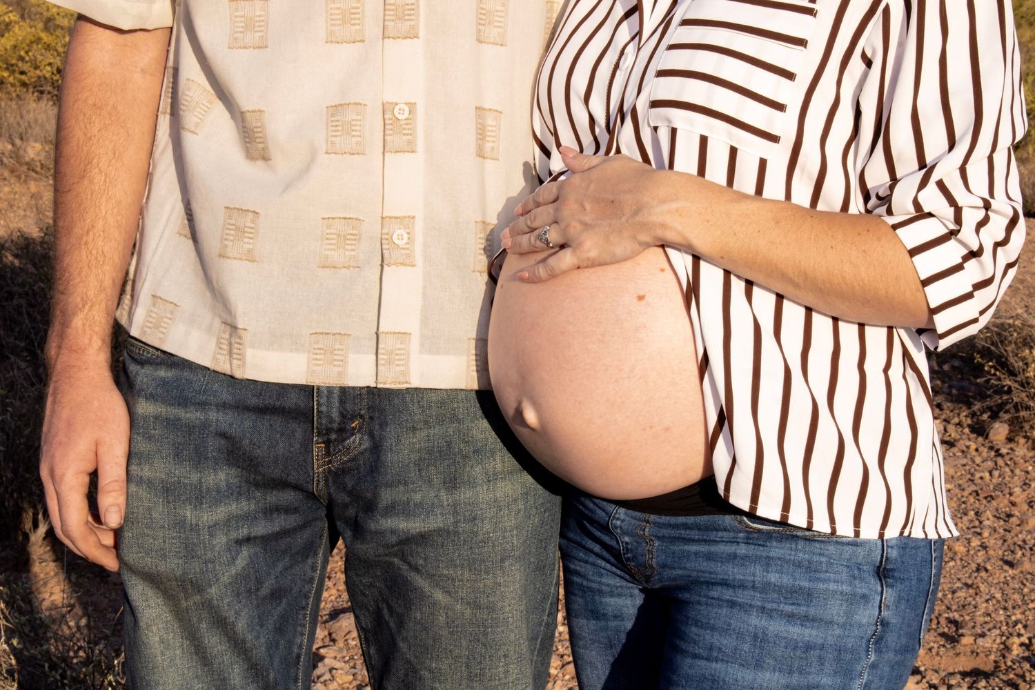 Couple posing outdoors showing pregnant belly, man in beige shirt and jeans, woman in striped top touching baby bump.