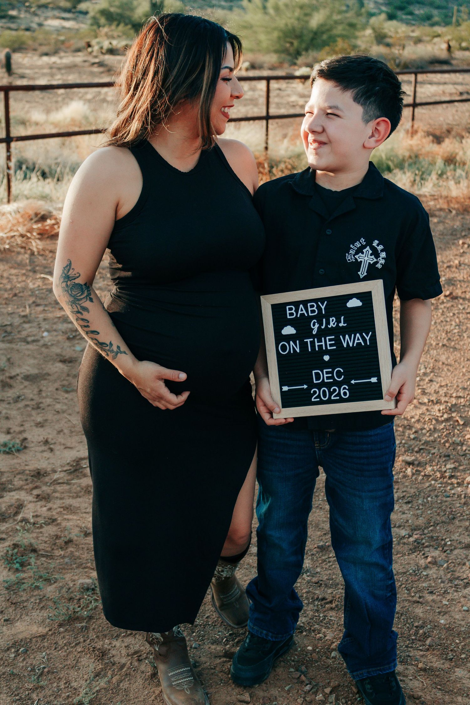 Pregnant woman in black dress with son holding baby girl announcement sign reading Dec 2026 outdoors.