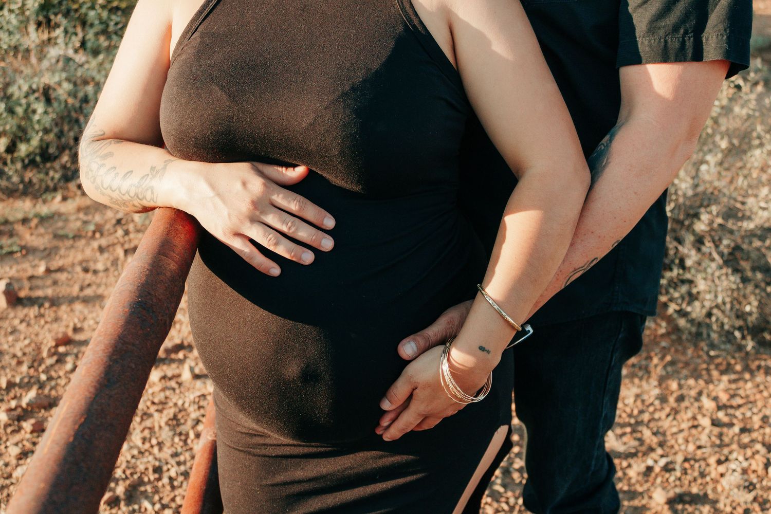 Pregnant woman in black dress with partner's hands on baby bump outdoors.