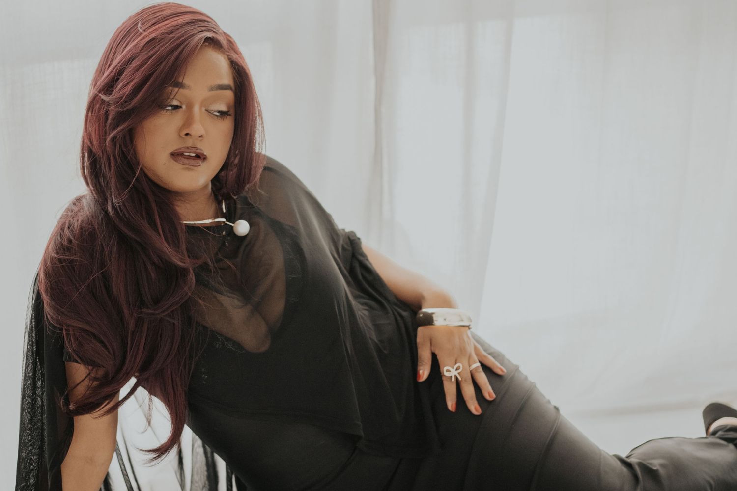 Woman with long red hair wearing a black top and leather pants, posing elegantly in a bright, airy room.