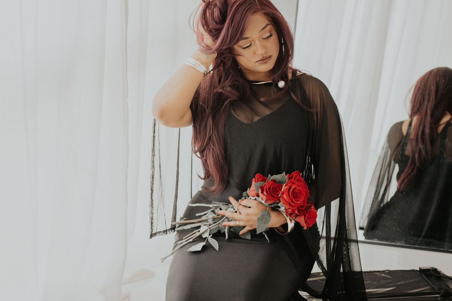 Woman in black dress holding red roses bouquet, with long red hair, posing elegantly near a mirror.