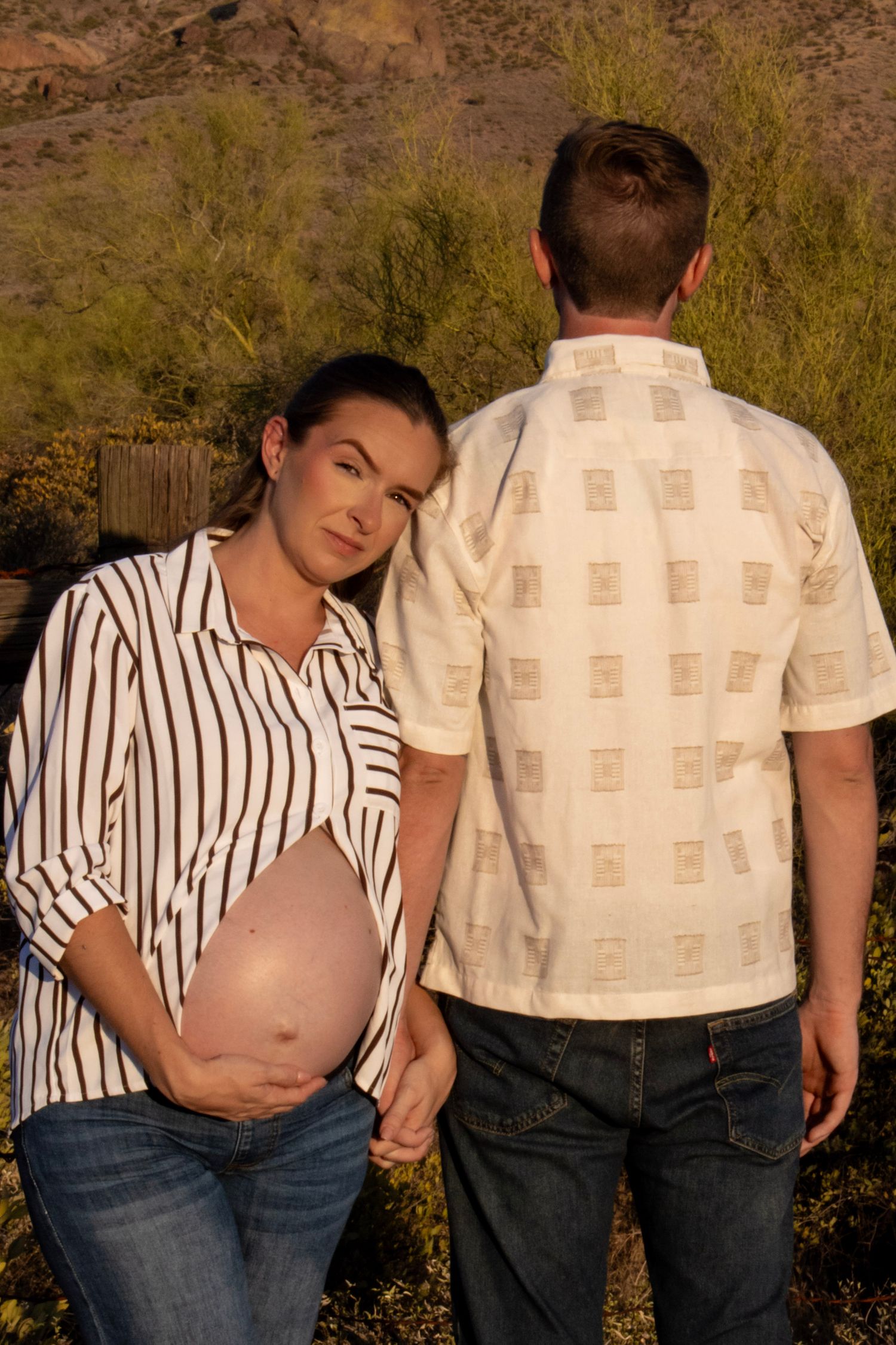 Pregnant woman in striped shirt holding partner's hand while showing baby bump in desert setting.