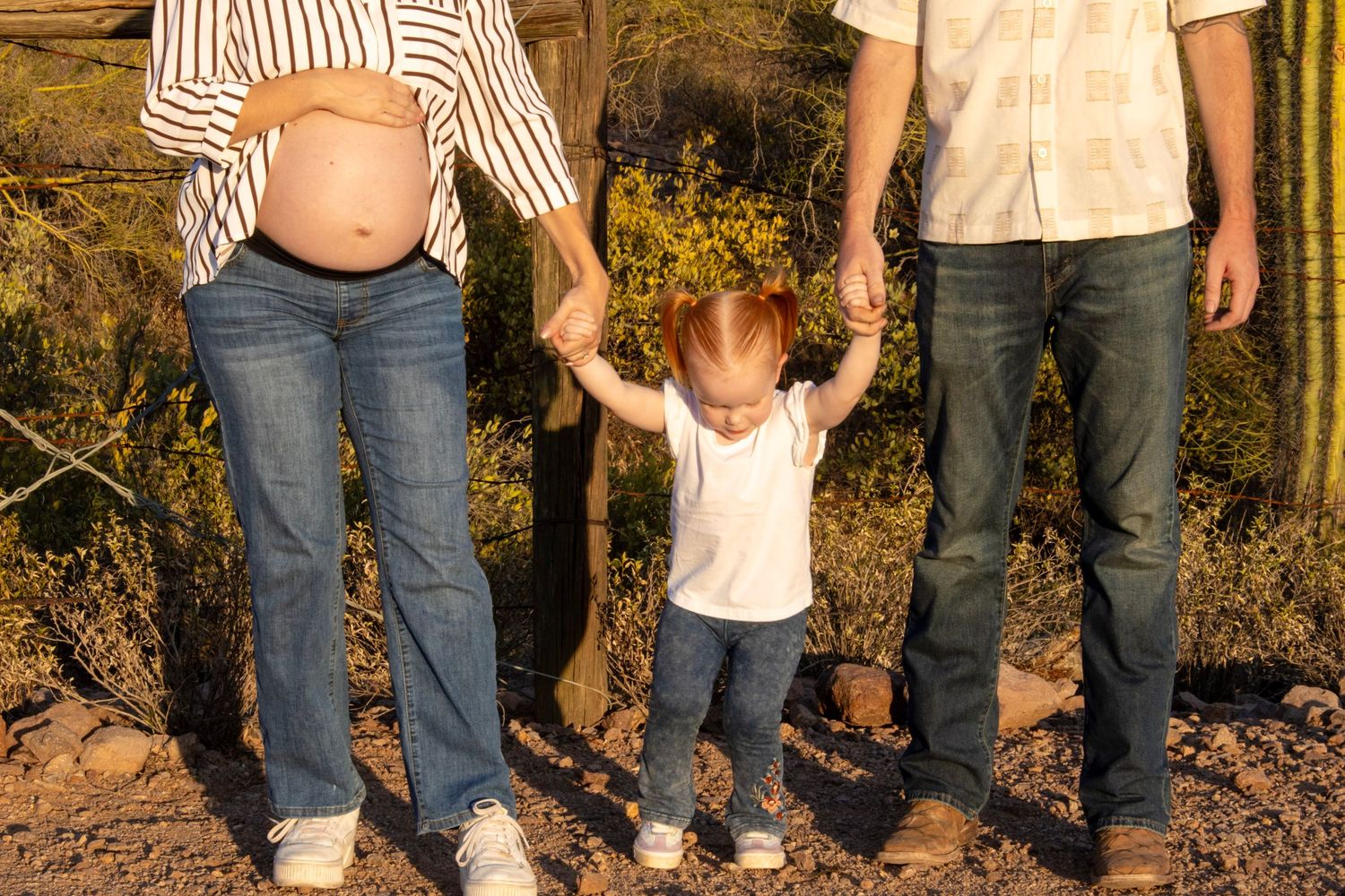 Pregnant mother and father holding toddler hands in sunny desert setting.