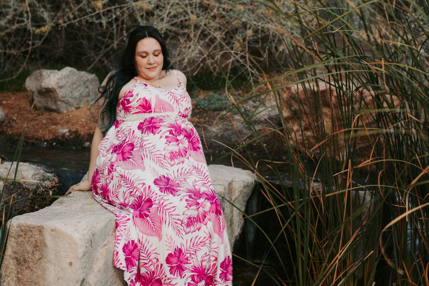 Pregnant woman in pink floral maxi dress sitting on rocks near a stream surrounded by tall grasses.