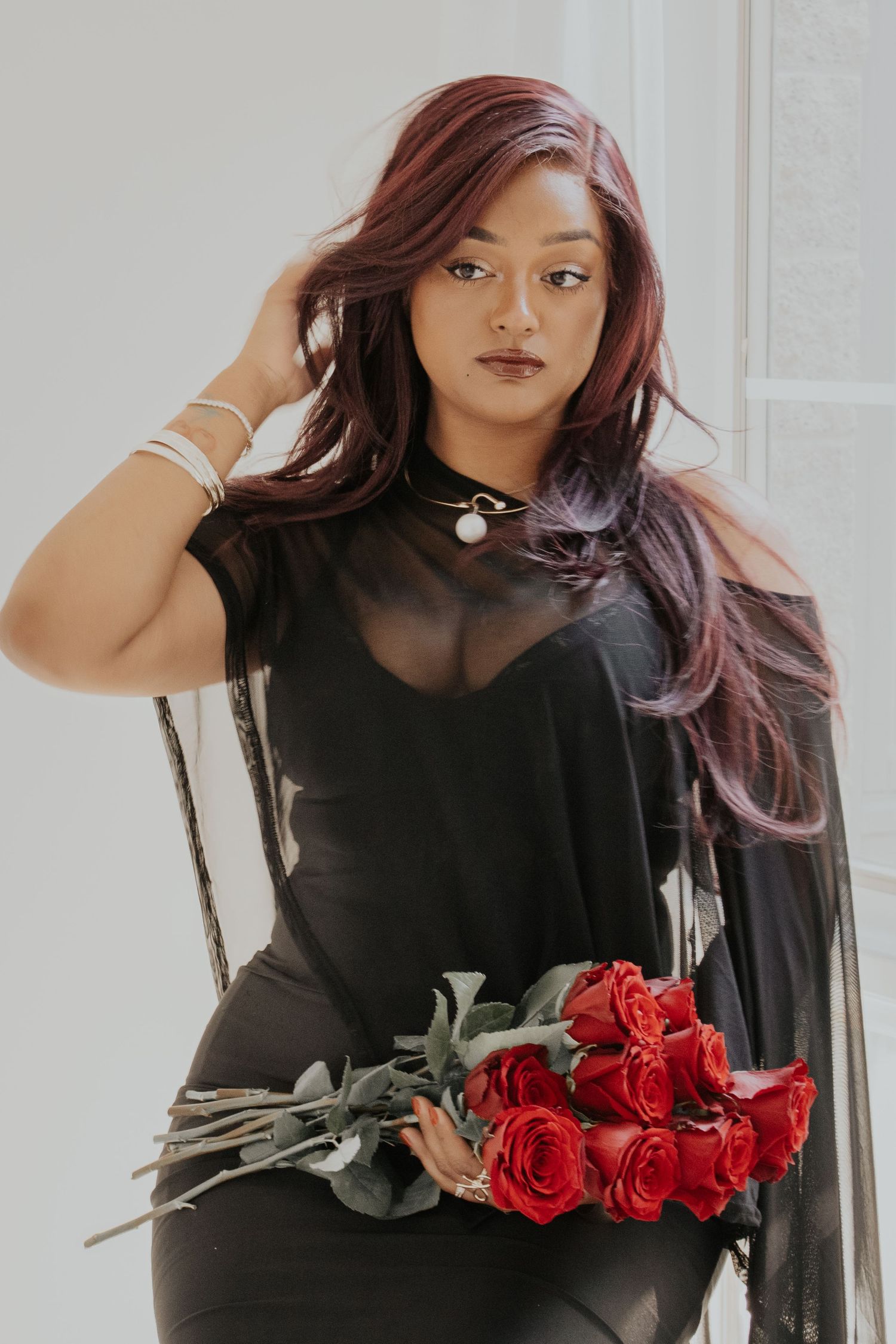 Woman with red-purple hair in black sheer dress holding red roses bouquet, posing elegantly indoors.