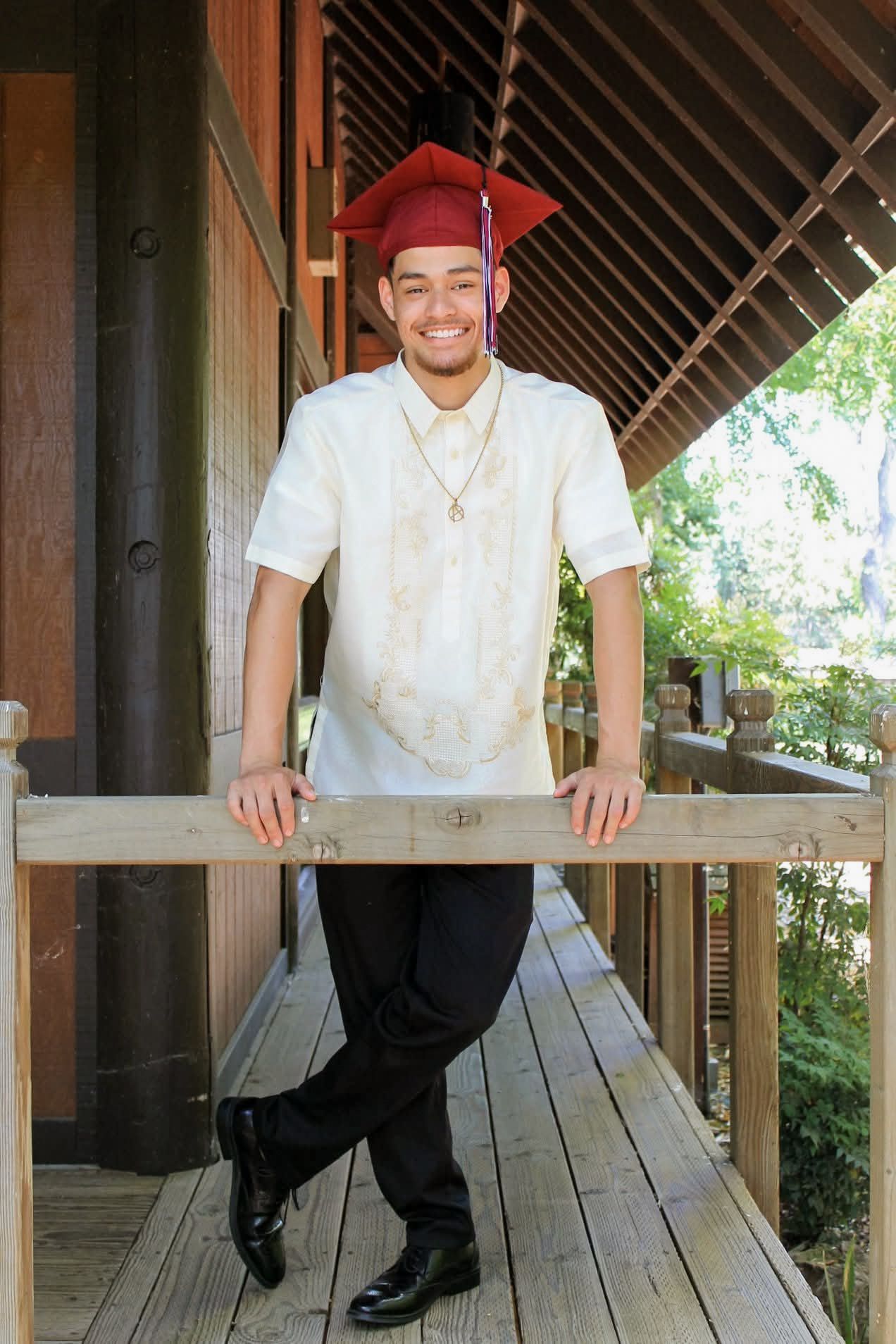 Young man in barong tagalog and red graduation cap smiling on wooden porch.