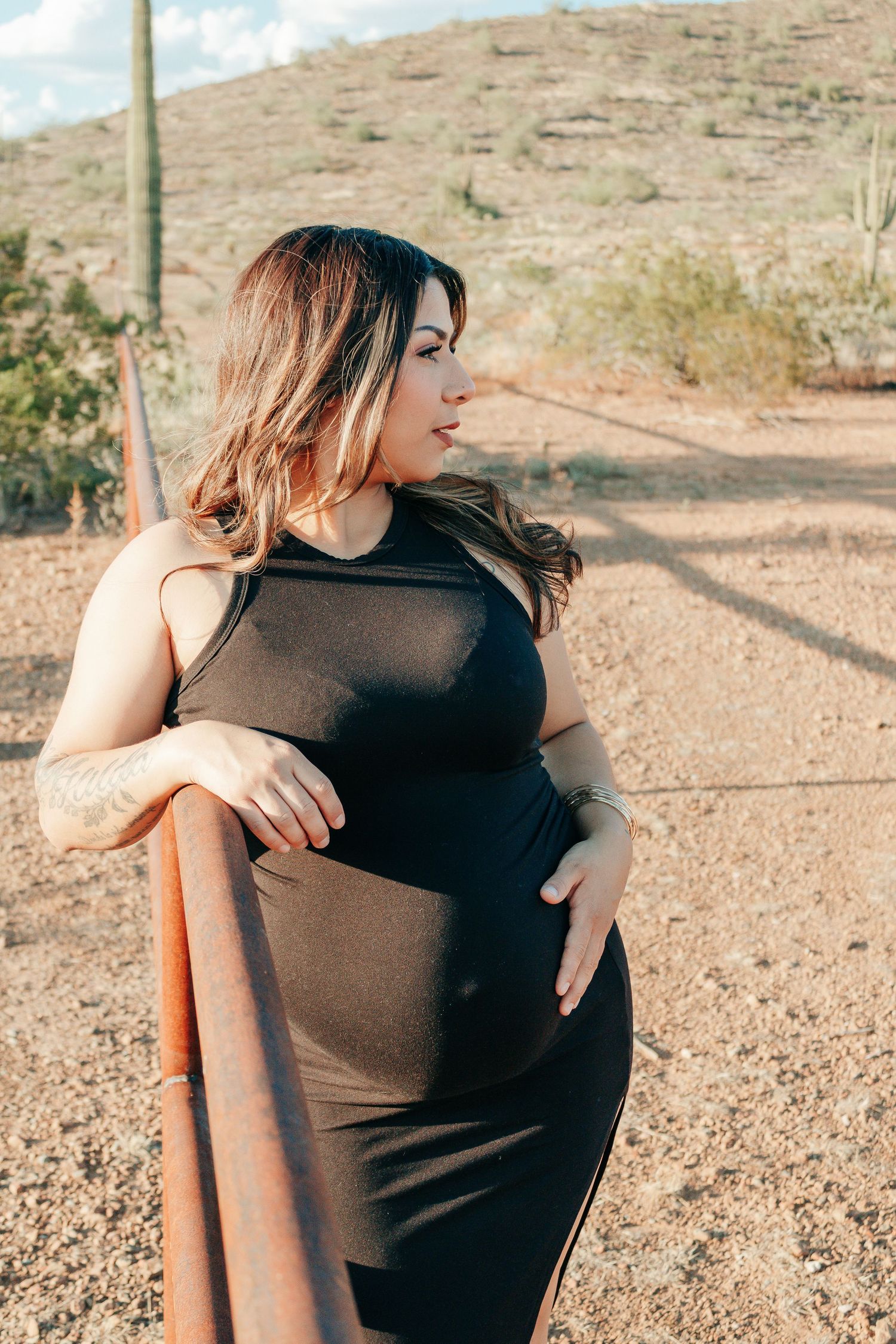 Pregnant woman in black dress leaning on rustic fence in desert landscape with mountains behind her.