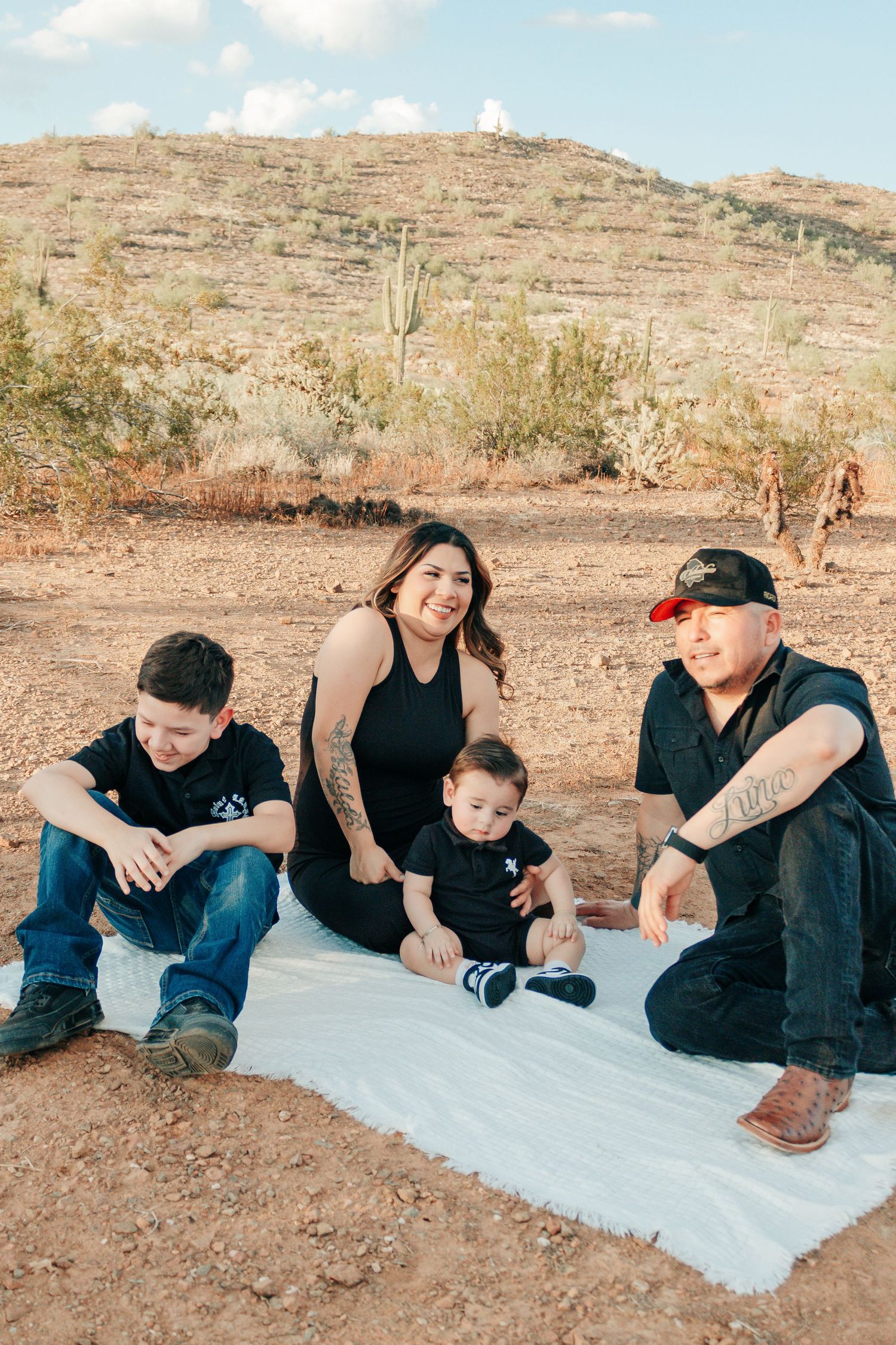 Family of four sitting on white blanket in desert landscape with saguaro cacti, all wearing matching black outfits.