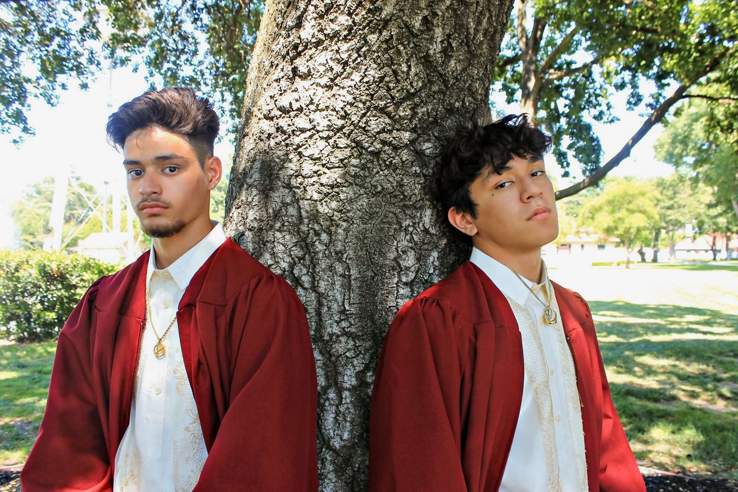 Two young men in red graduation gowns leaning against a tree trunk in a sunny park.
