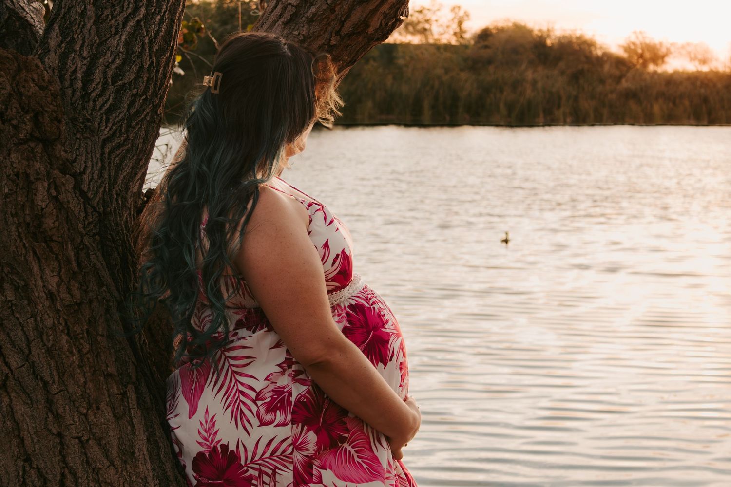 Pregnant woman in floral dress leaning against tree, gazing at serene lake during golden hour sunset.
