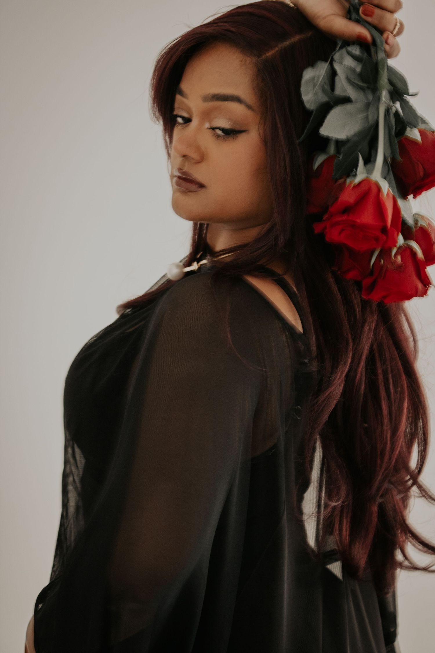 A young woman with long red-brown hair in a black sheer top holds red roses, posing elegantly against a light background.