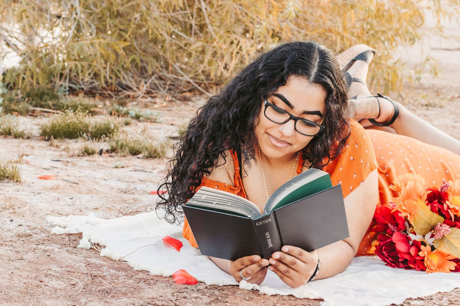 Young woman with curly hair and glasses reading a book outdoors while lying on a blanket in an orange dress.