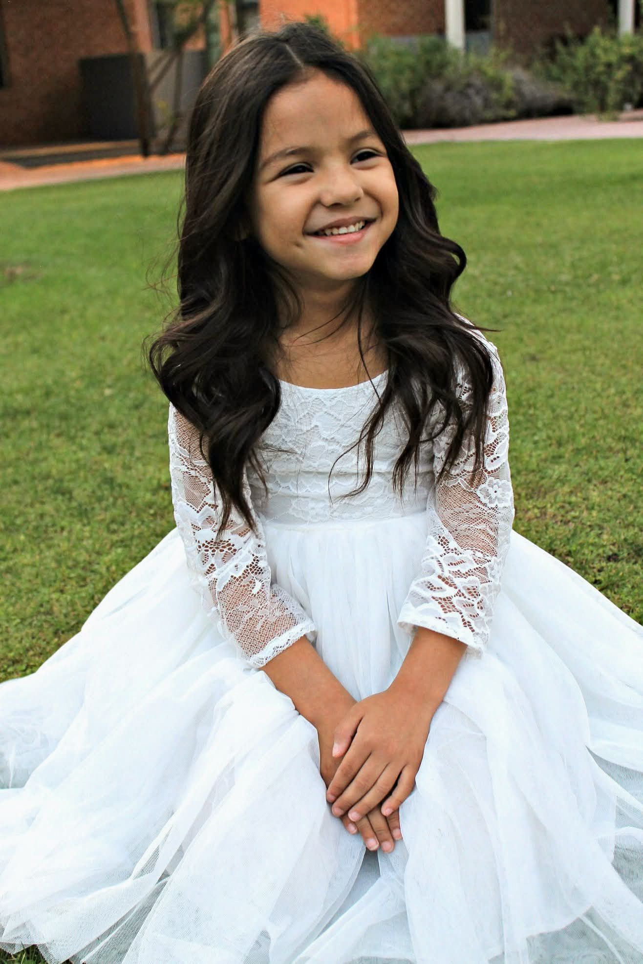 Smiling young girl in white lace dress sits on grass in sunny garden.