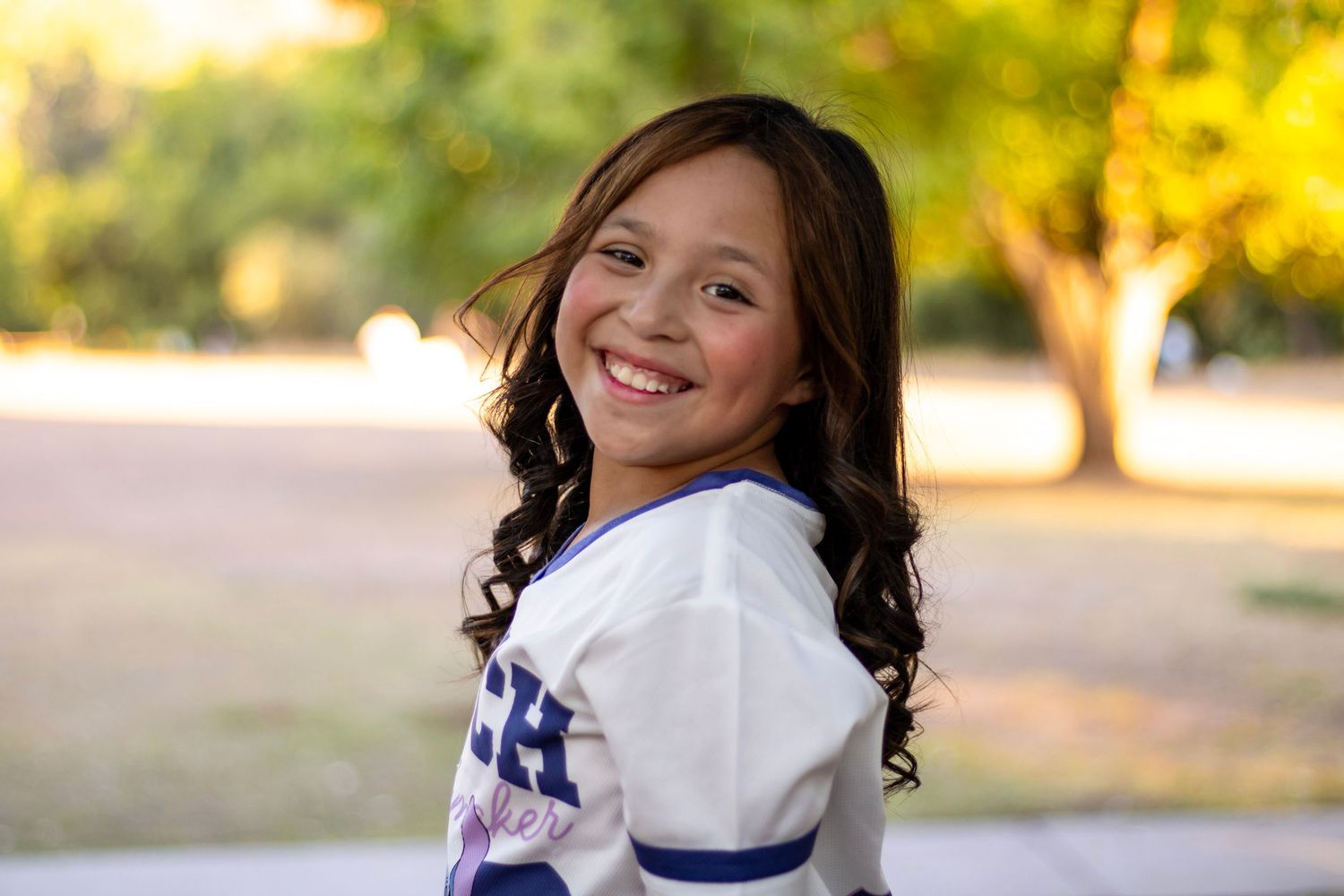 Smiling young girl with curly dark hair wearing a white sports jersey in a sunny park setting.