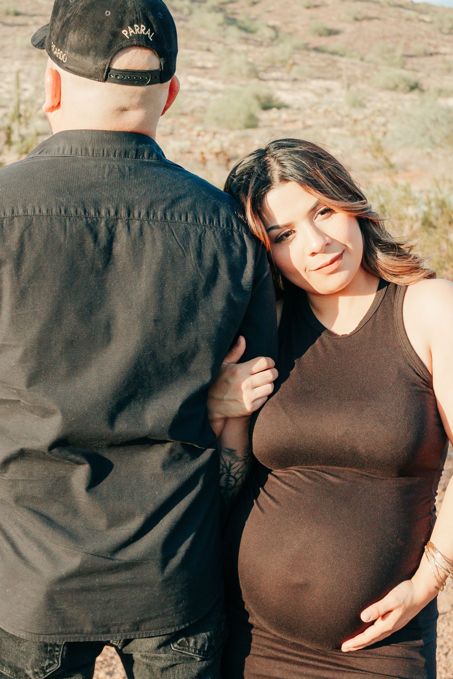 Pregnant woman in brown dress smiles outdoors with partner in black shirt, cradling baby bump in desert landscape.