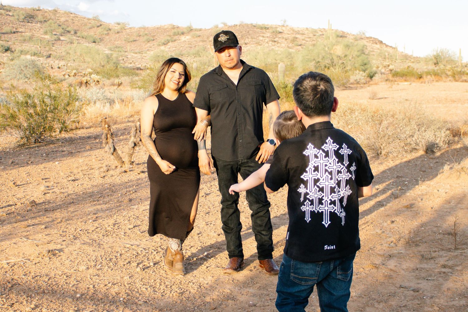 Pregnant woman in black dress with partner and child in desert landscape during golden hour maternity photo session.