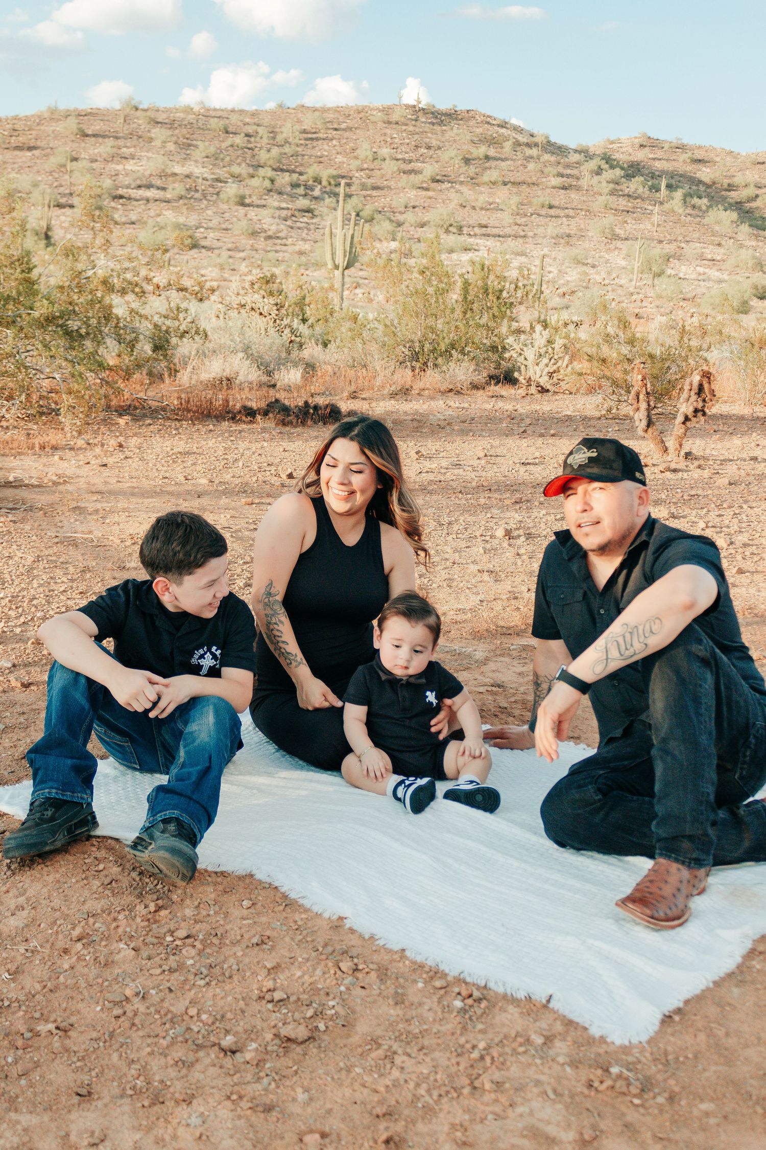 Family of four sitting on white blanket in desert landscape with saguaro cactus and hill backdrop.