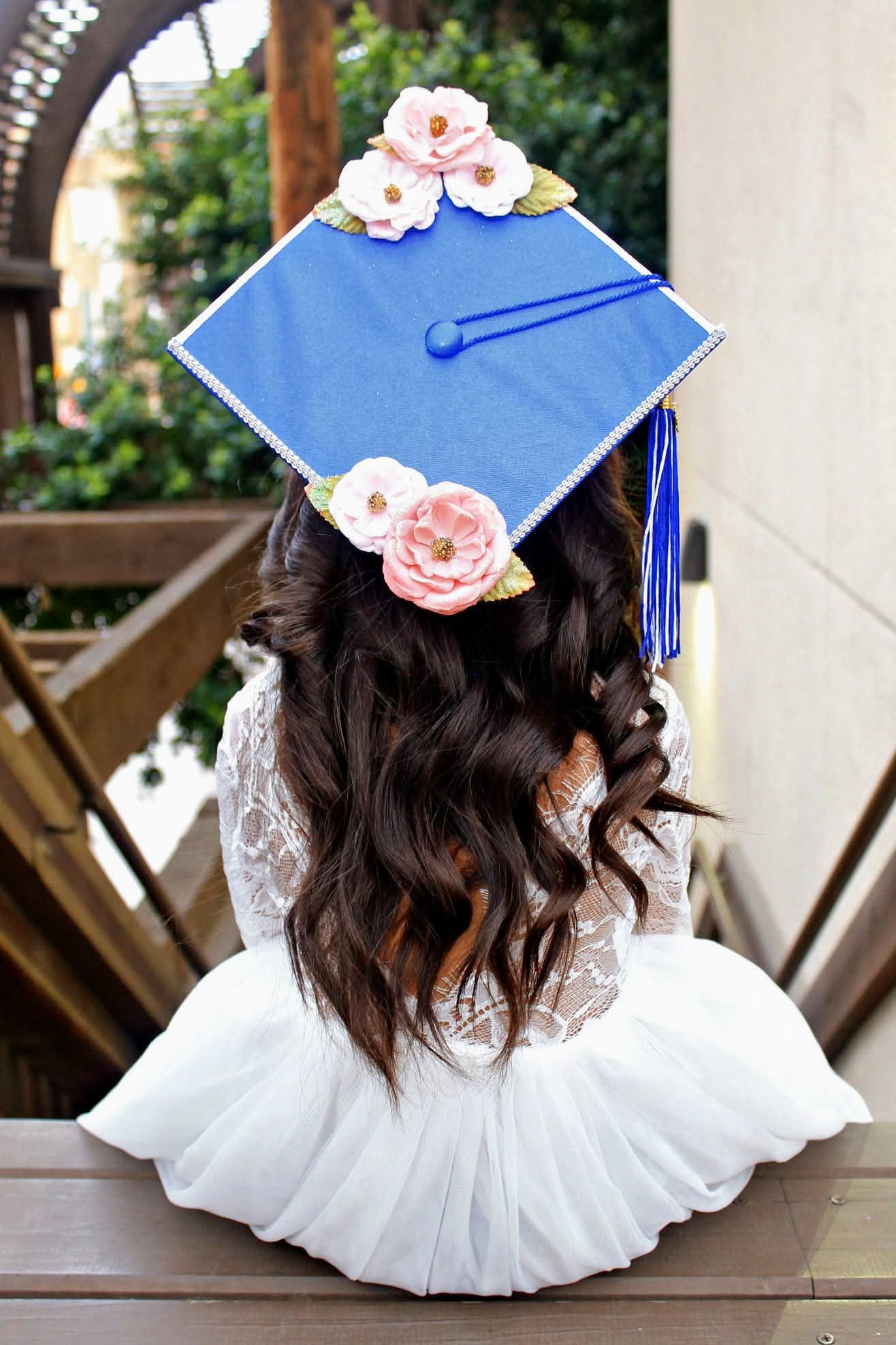 Graduate with decorated blue mortarboard cap adorned with pink flowers and long wavy brown hair.