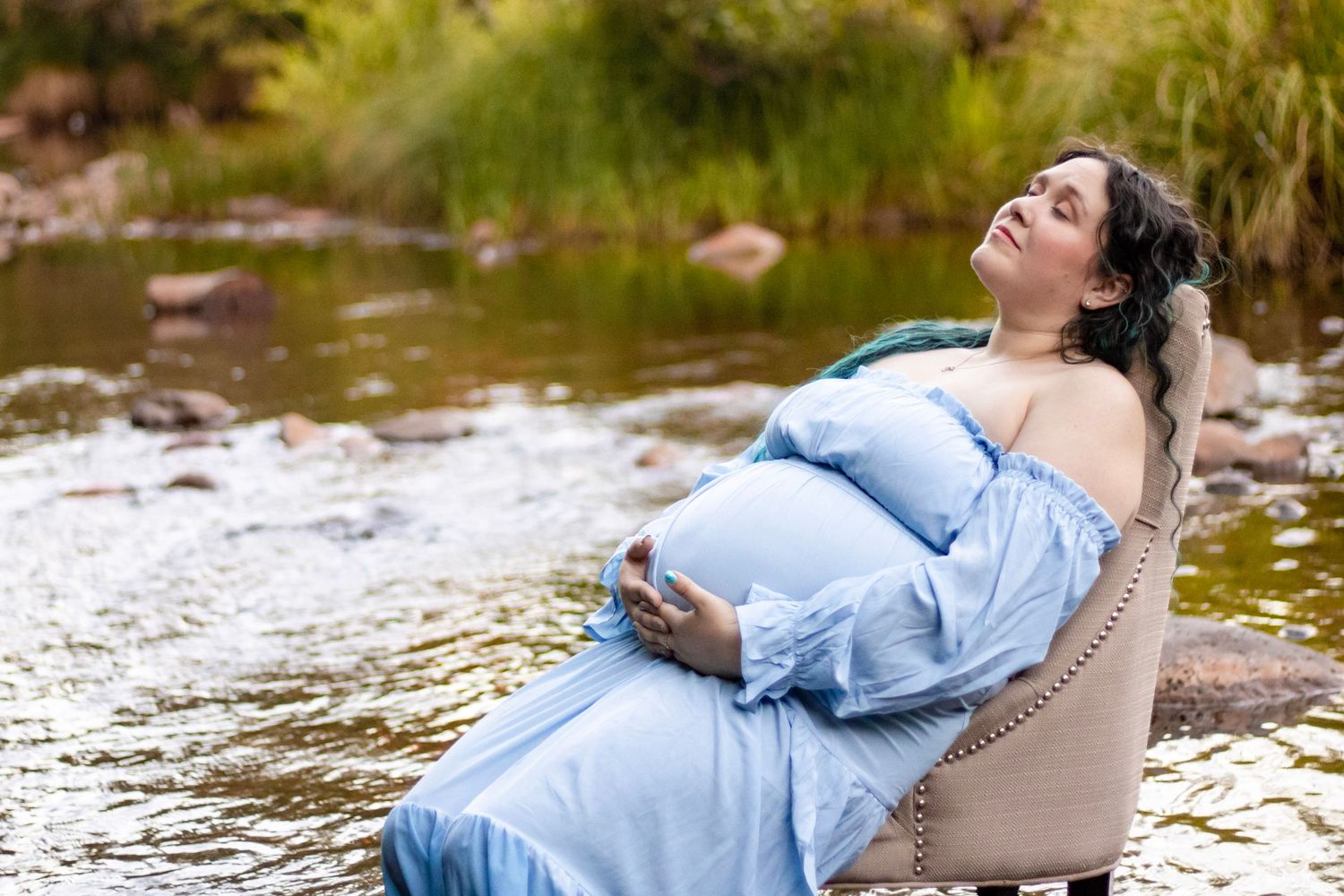 Pregnant woman in blue gown relaxing in armchair beside a peaceful rocky river.