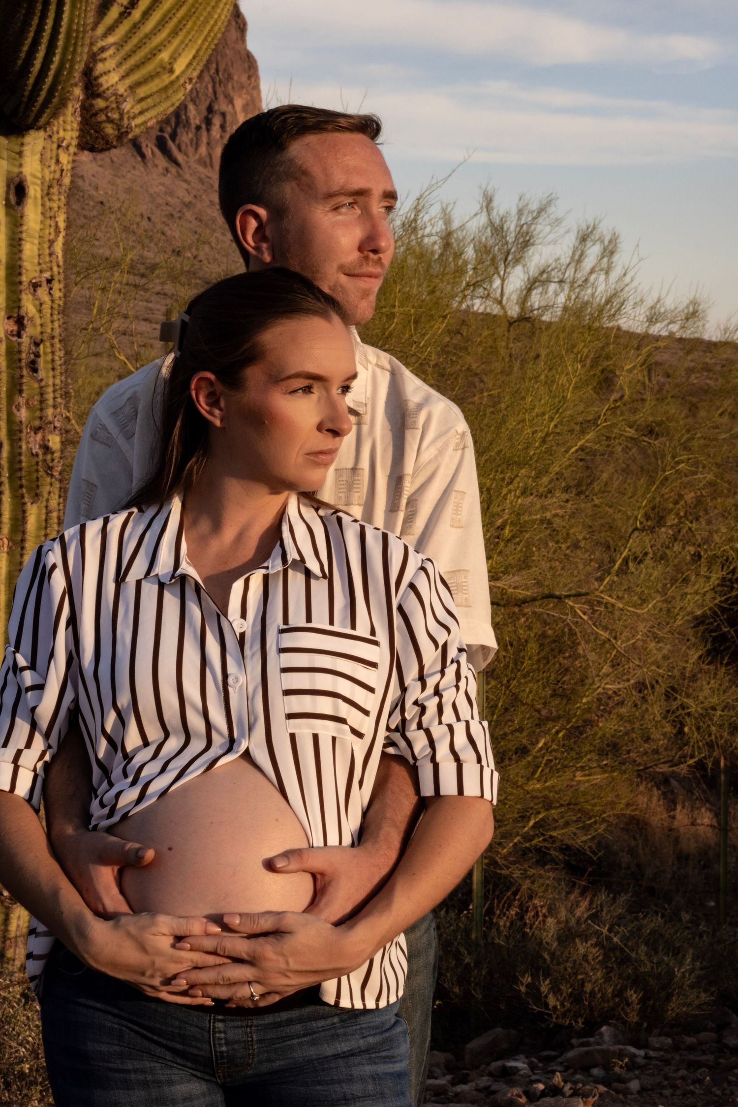 Couple embracing pregnant belly together near saguaro cactus in golden Arizona desert light.