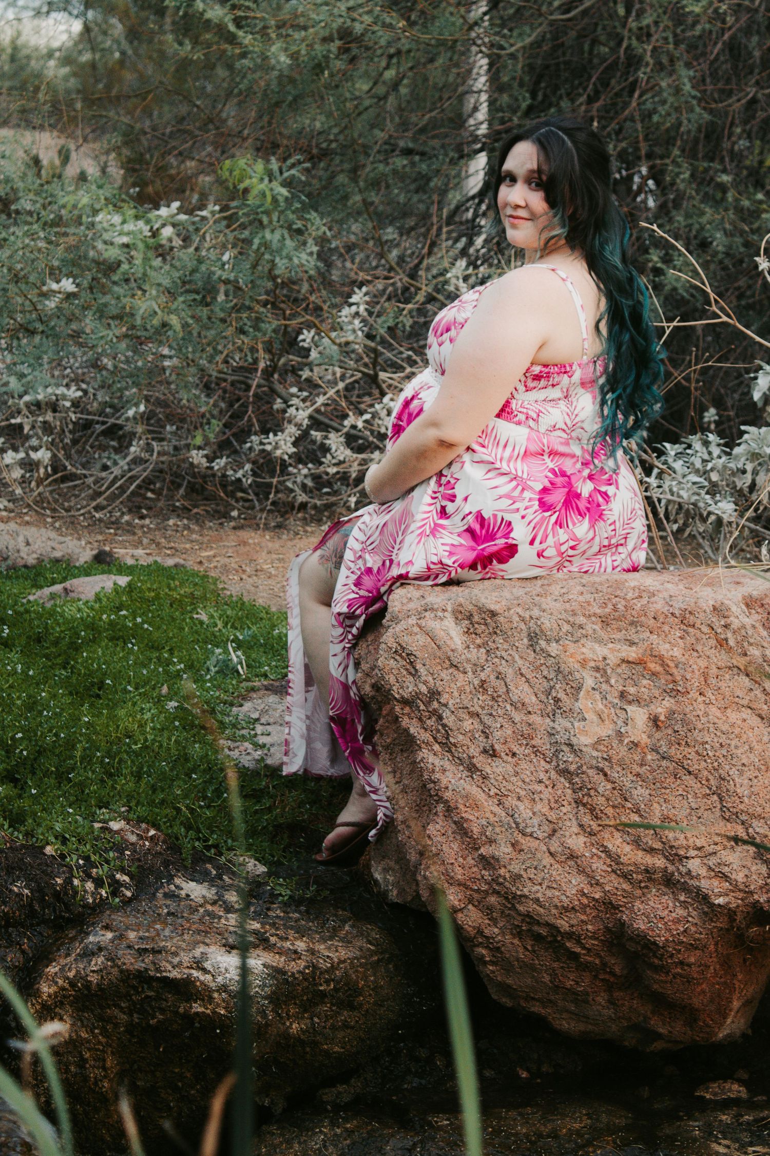 Woman in pink floral dress sitting on a large rock in a natural outdoor setting with lush greenery.