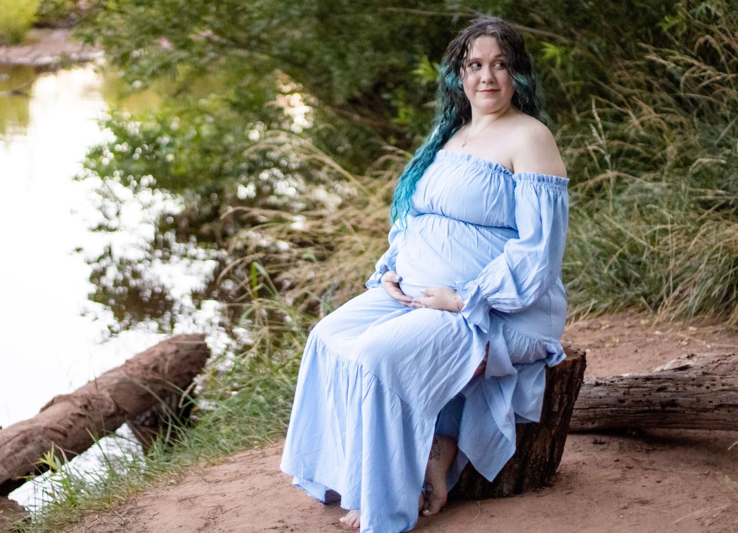 Pregnant woman in a blue off-shoulder maxi dress sitting on a log near a river surrounded by greenery.