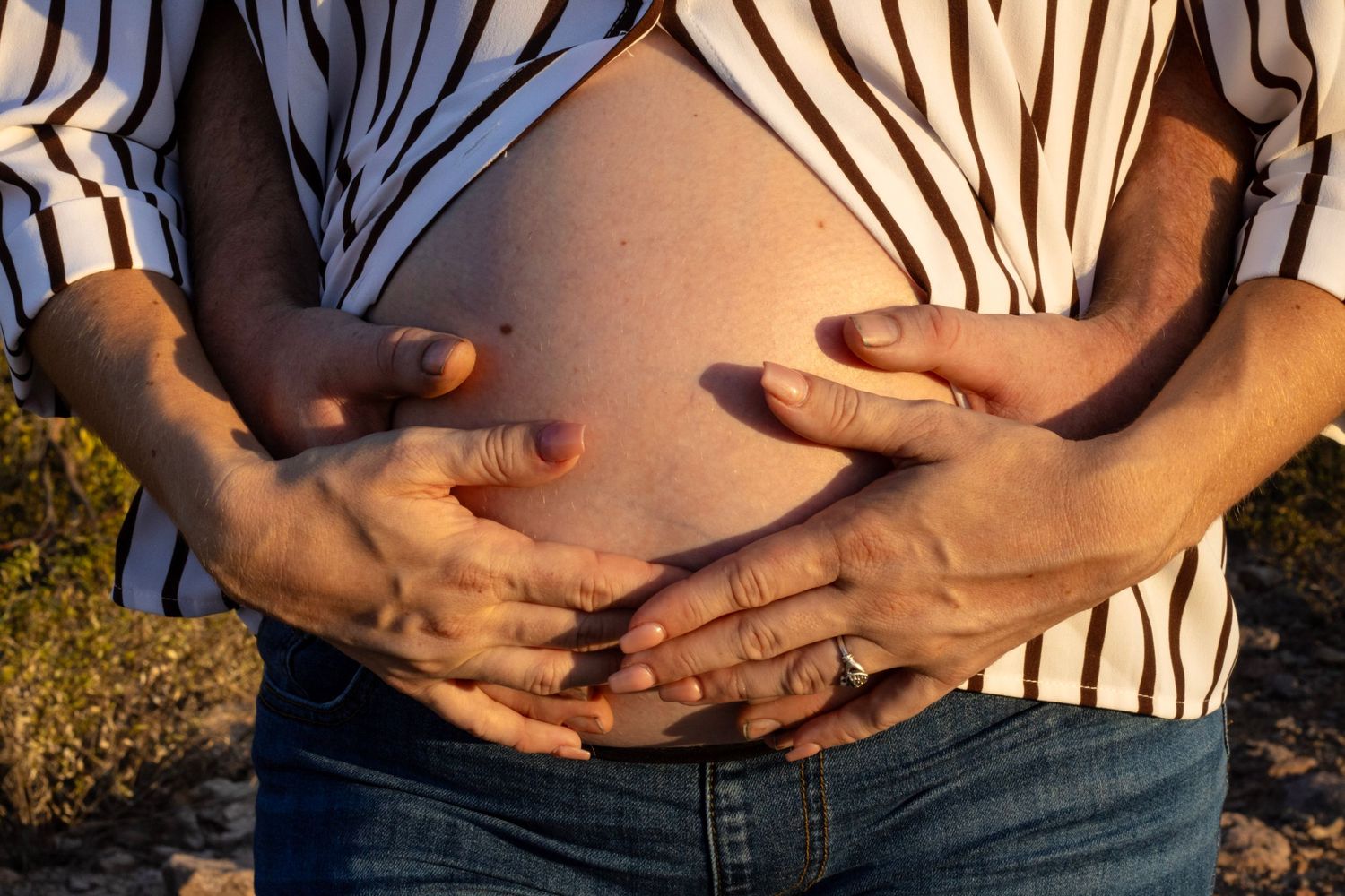 Two pairs of hands gently cradling pregnant belly in striped shirt during outdoor maternity session.