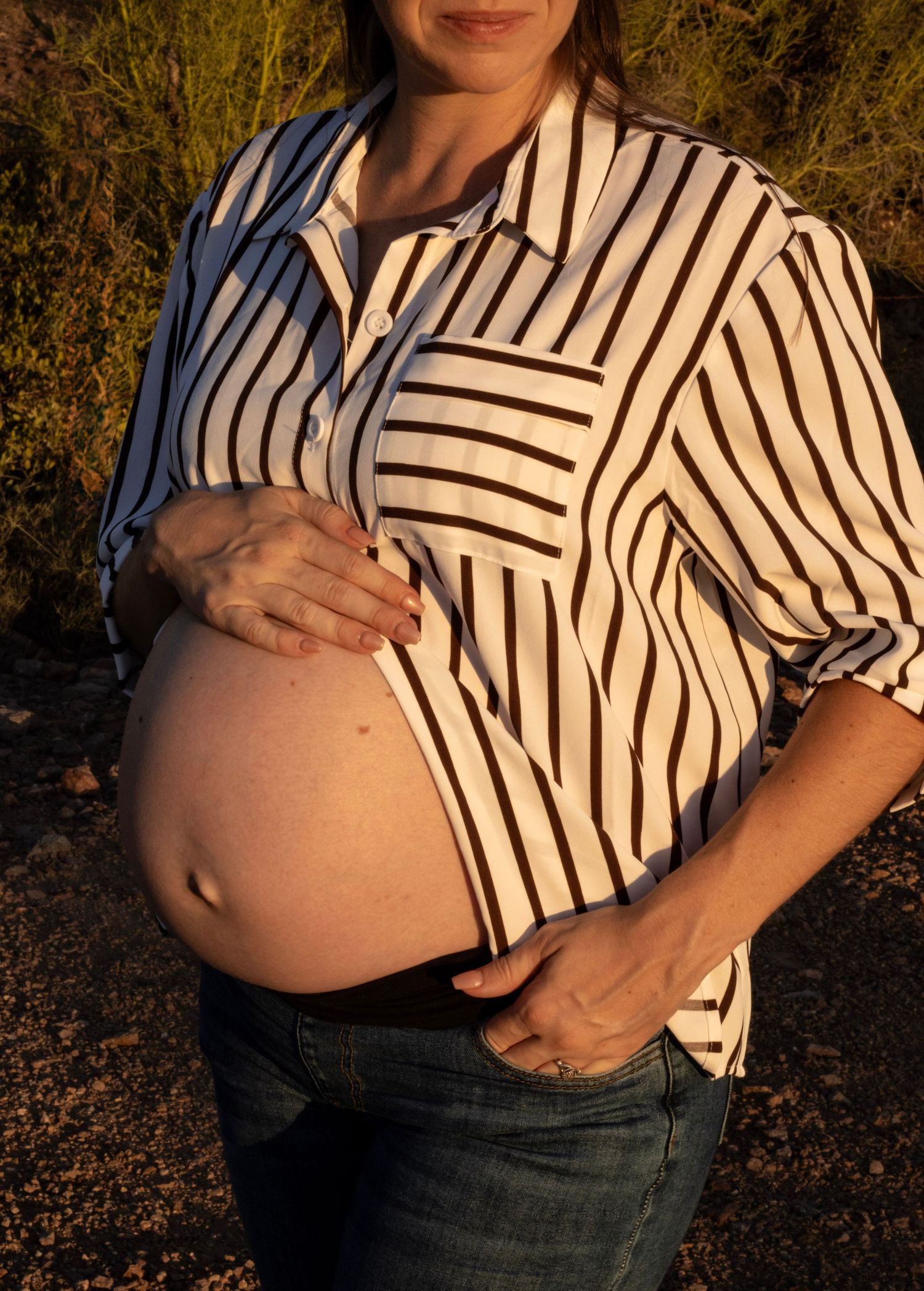 Close-up of pregnant woman in striped shirt cradling bare baby bump in desert maternity photo.