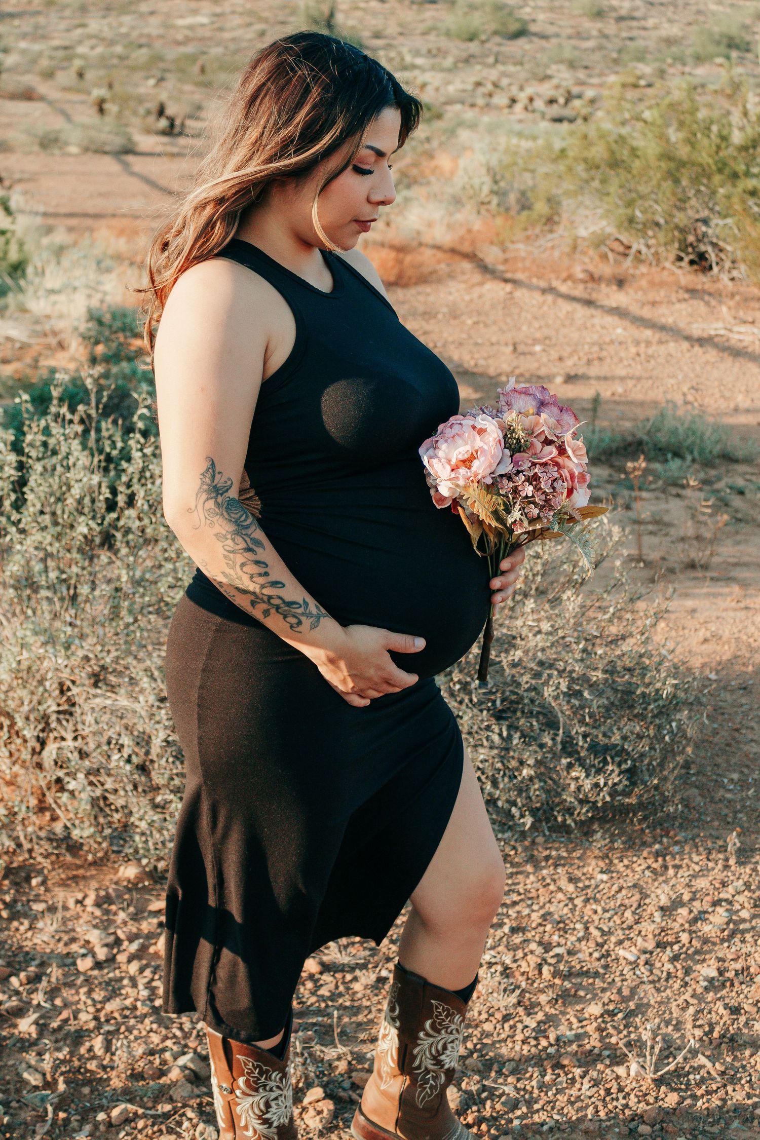 Pregnant woman in black dress holding pink flower bouquet, showing baby bump outdoors in desert setting with cowboy boots.