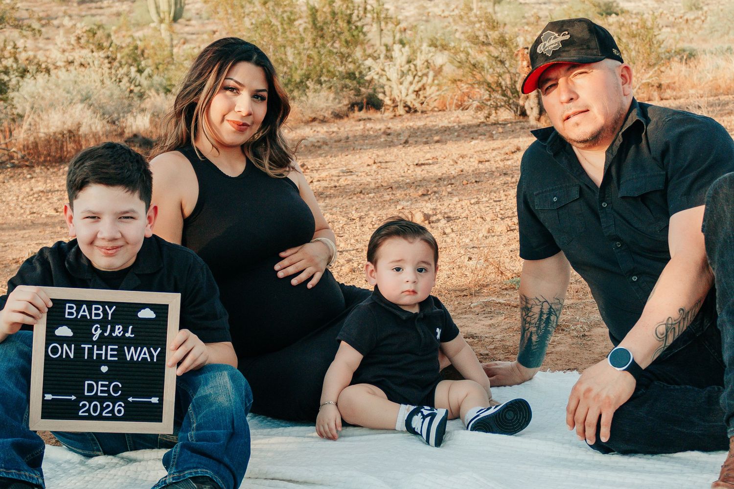 Family pregnancy announcement photo with letterboard sign reading Baby Girl On The Way Dec 2026, outdoors in desert setting.