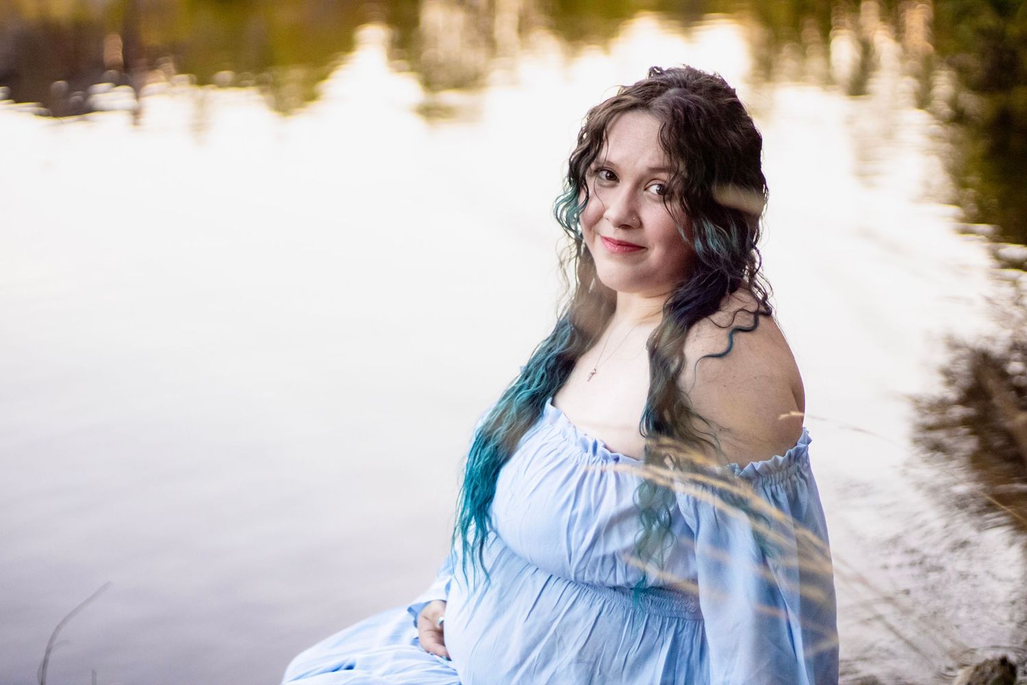 Pregnant woman in blue off-shoulder gown sitting by reflective lake water in a dreamy maternity photo.