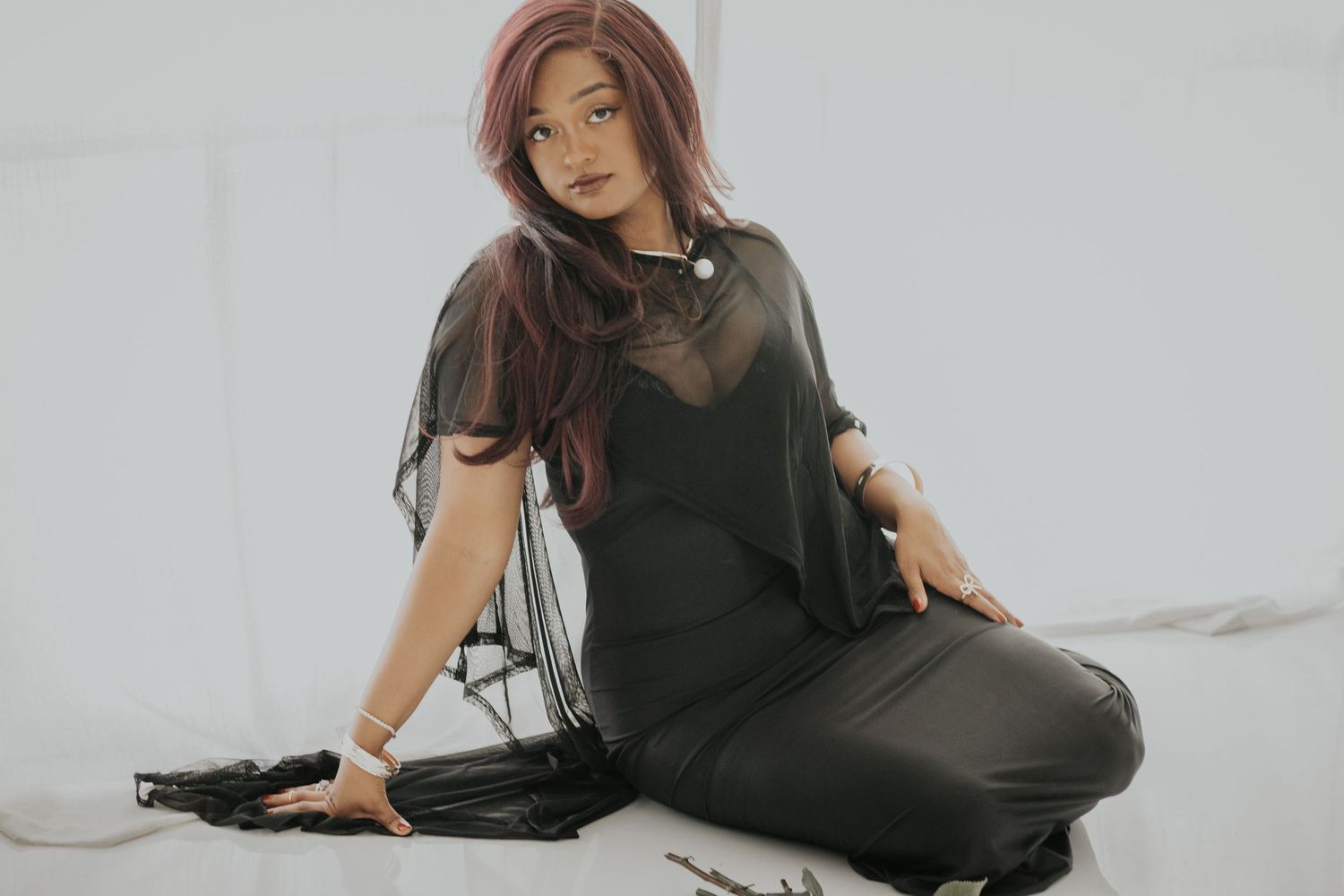 Woman with red hair poses in black sheer top and leather pants against white background.