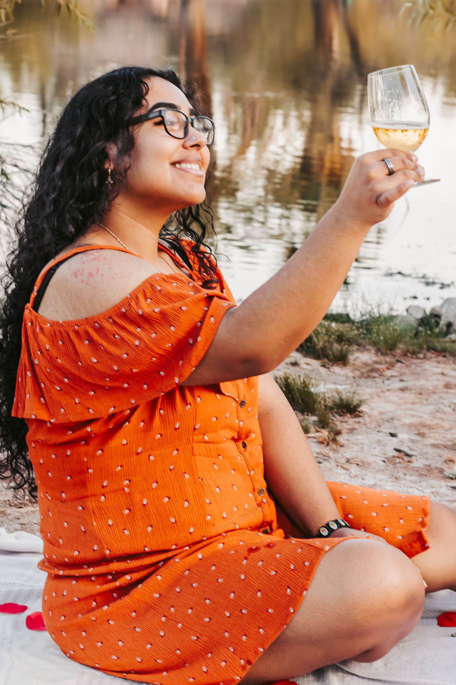 Woman in orange polka dot dress sitting outdoors, raising a glass of white wine near a lake, smiling joyfully.