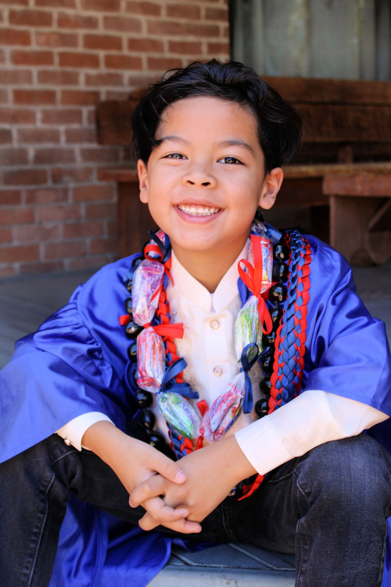 Smiling boy in blue graduation gown with colorful candy lei sitting outdoors.