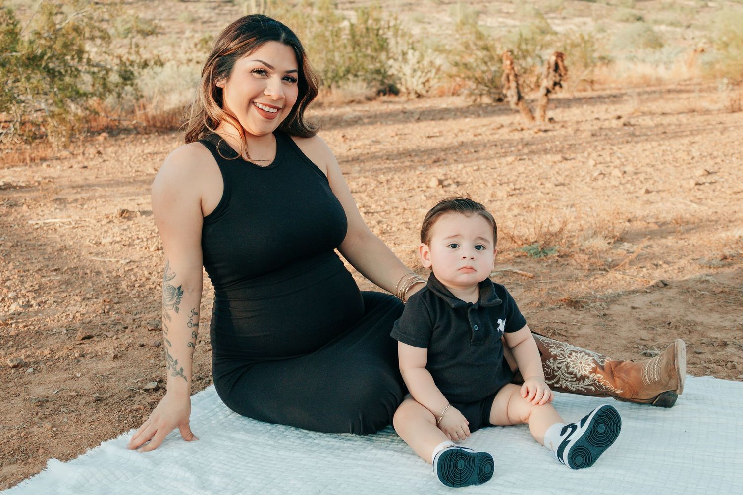 Pregnant woman in black dress sits outdoors on white blanket with baby boy in black outfit, desert landscape background.