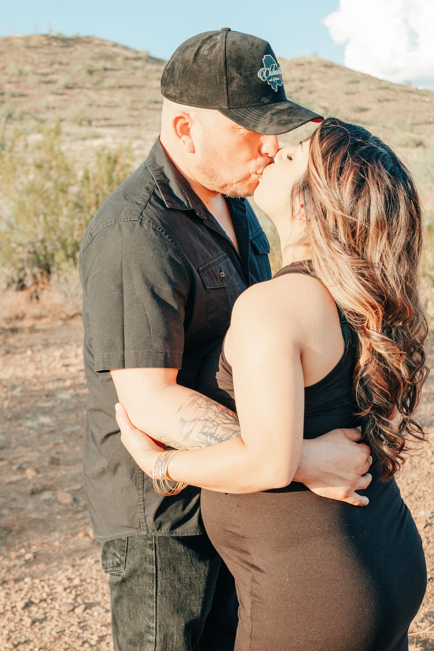 Couple sharing a romantic kiss outdoors in a desert landscape, man in black cap and shirt, woman in dark dress.