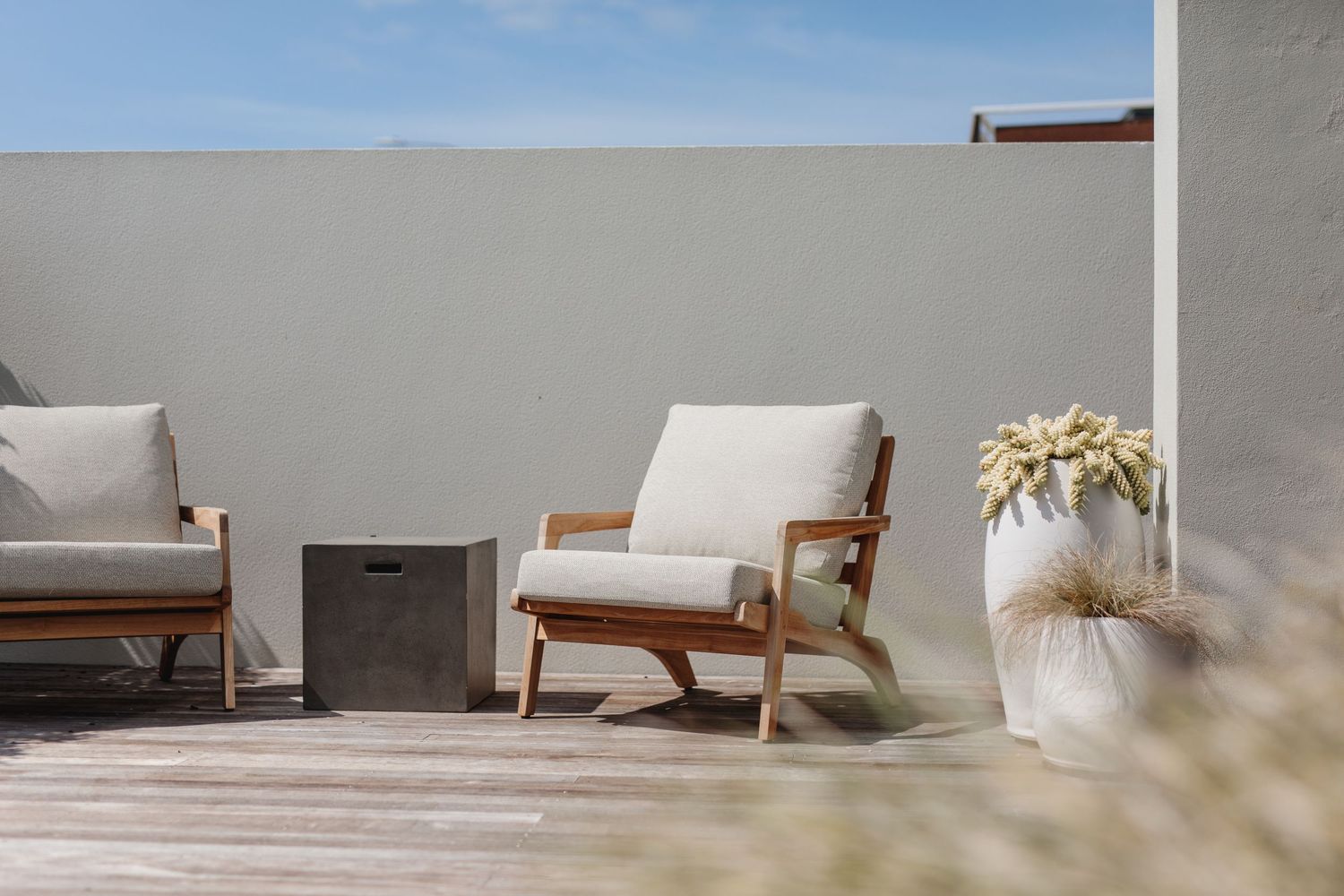 Minimalist patio setup with wooden lounge chairs and white cushions against a light colored wall.