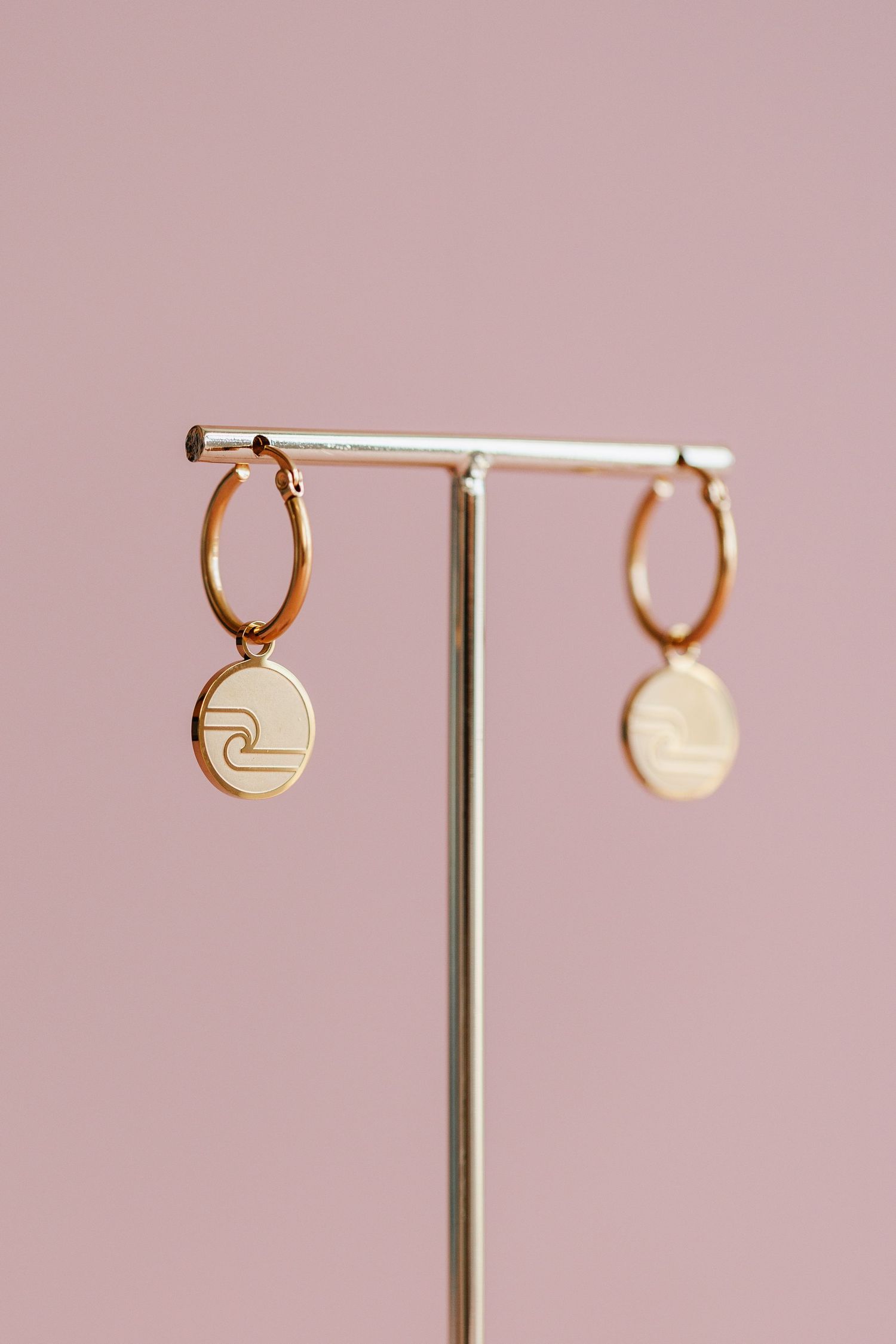 Gold hoop earrings with circular pendant charms displayed on a minimalist jewelry stand against pink background.