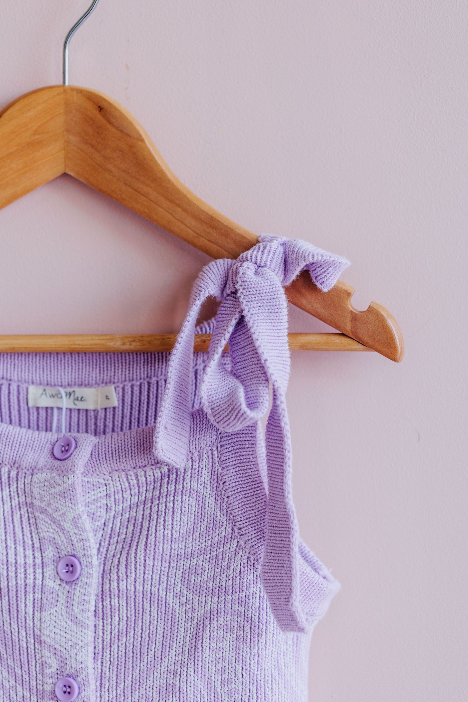 Lavender knit tank top with buttons hanging on wooden hanger against pink wall.