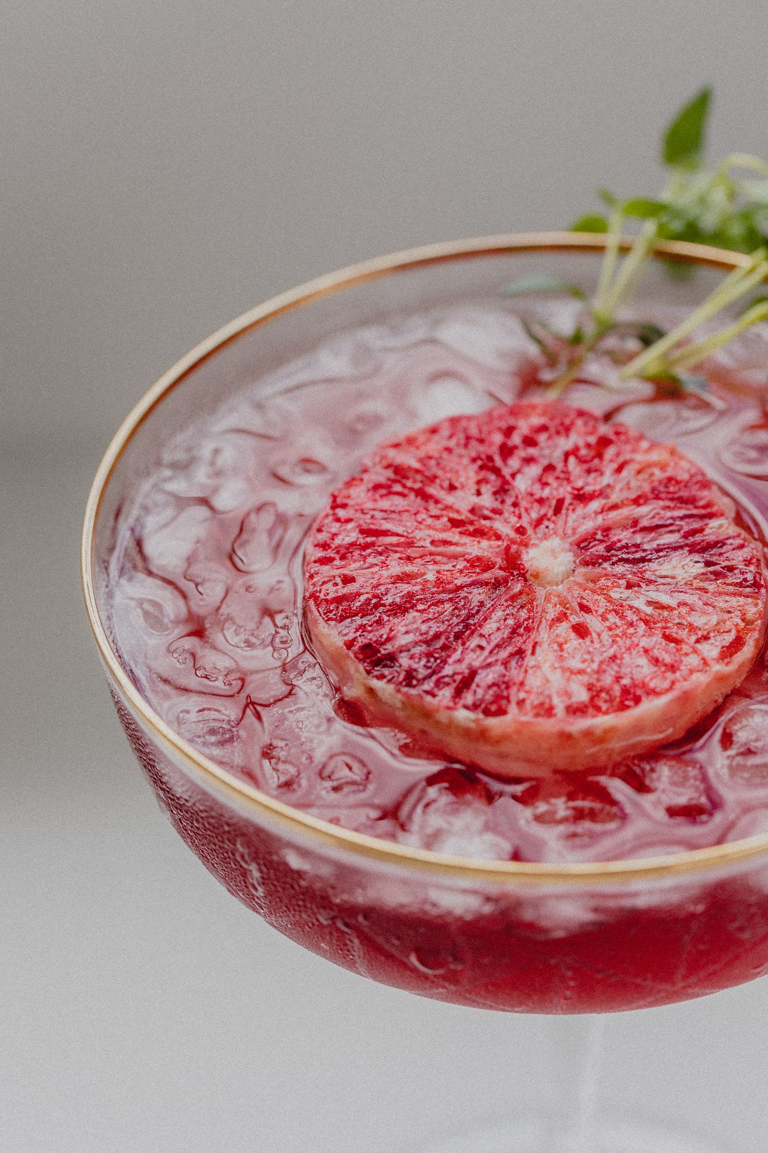 Pink grapefruit cocktail garnished with herbs and ice in a coupe glass.